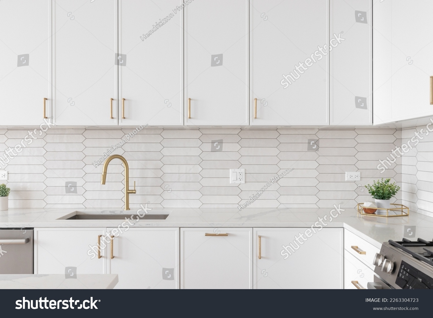 A beautiful kitchen faucet detail with white cabinets, a gold faucet, white marble countertops, and a brown picket ceramic tile backsplash. #2263304723