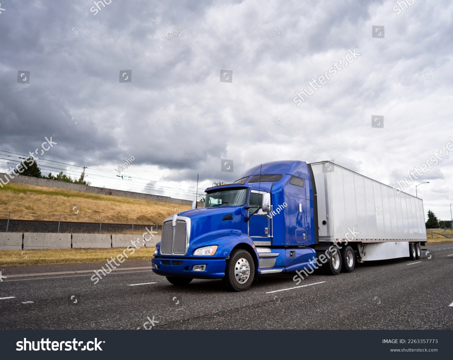 Classic dark blue big rig American popular bonnet semi truck with dry van semi trailer going on wide highway with stormy sky caring commercial cargo for delivery to business warehouse  #2263357773