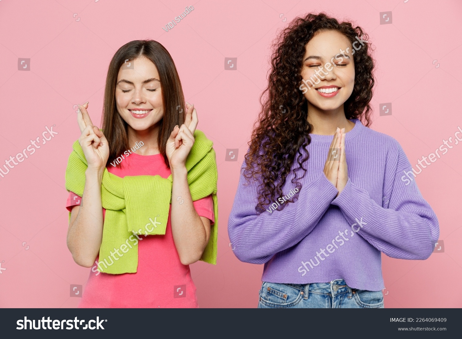 Young two friends women wears green purple shirts looking camera together wait for special moment, keep fingers crossed, hands folded in prayer gesture isolated on pastel plain light pink background #2264069409