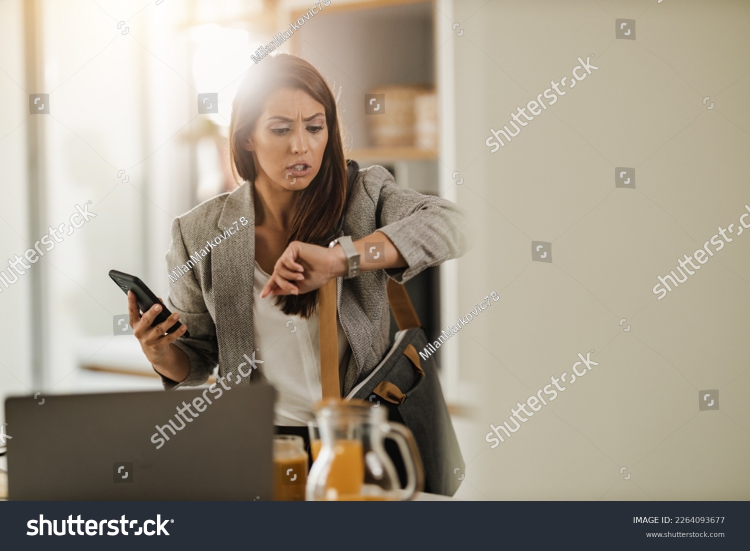 Shot of a multi-tasking young business woman looking on watch and using smartphone in her kitchen while getting ready to go to work. #2264093677