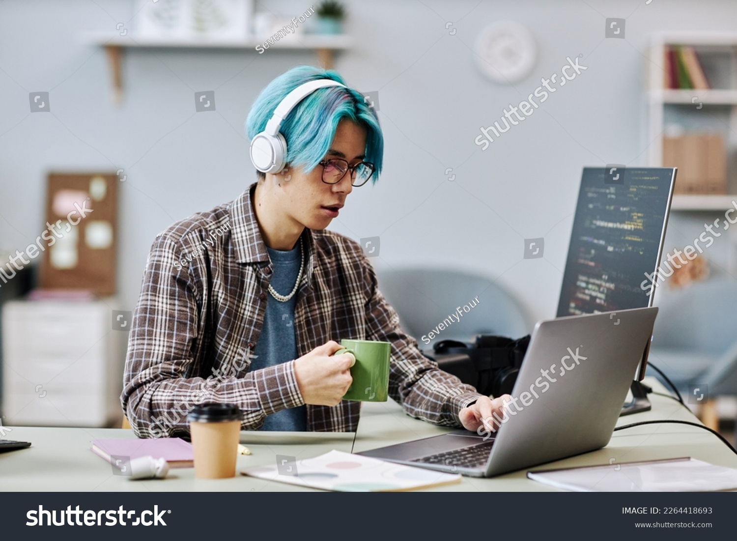 Portrait of young man with blue hair using computer in office and wearing headphones while writing code #2264418693
