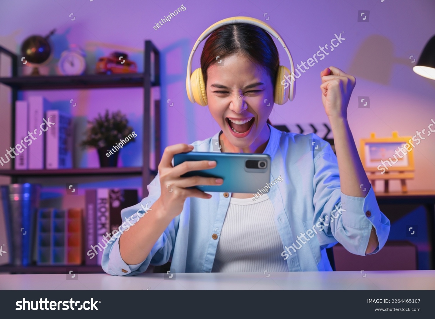 Excited Young Asian woman wearing headset and playing online game on smartphone at night modern house. #2264465107
