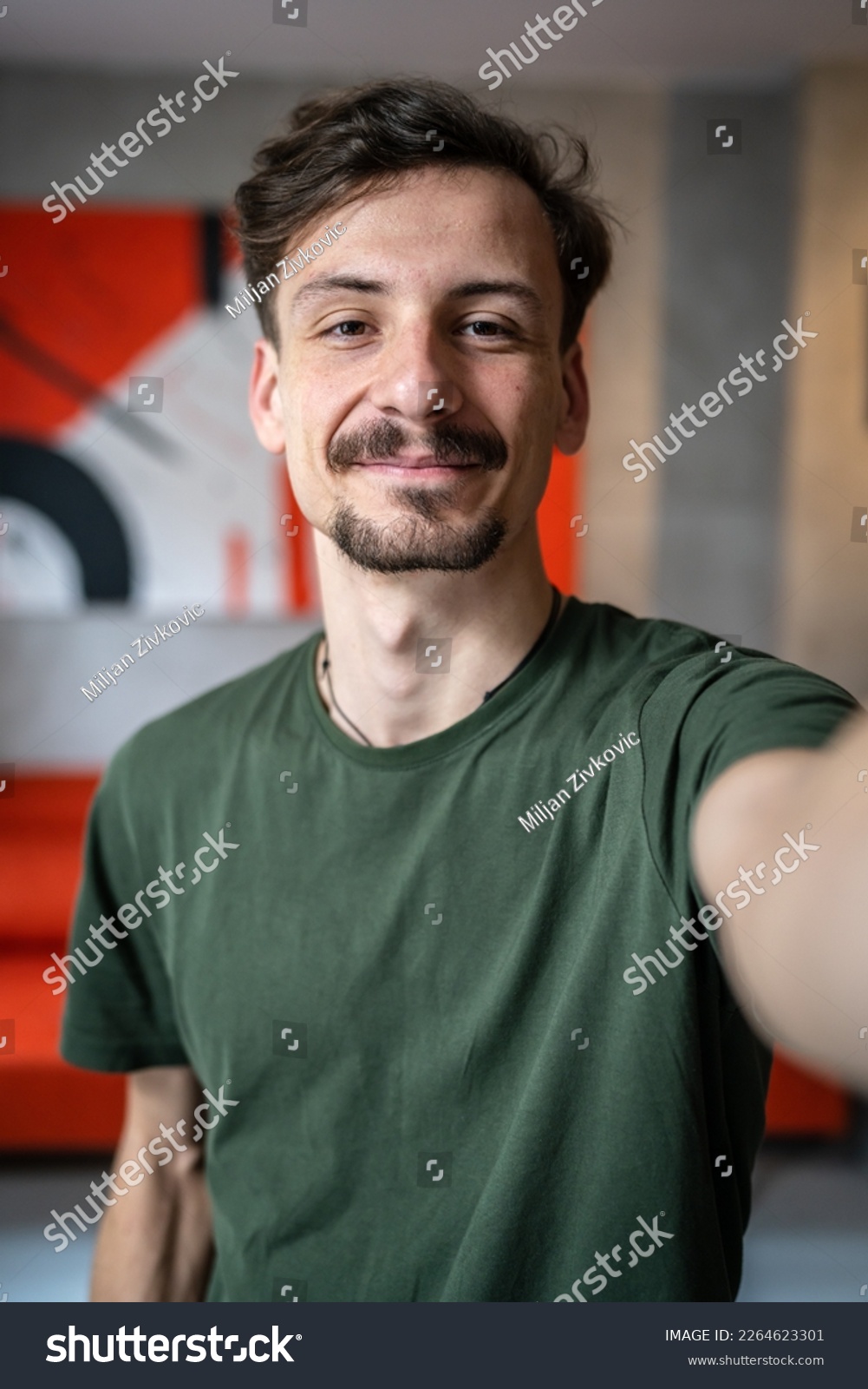 One man young adult caucasian male standing at home with brown hair mustaches and beard looking to the camera happy smile confident real people green t shirt UGC selfie user generated content #2264623301