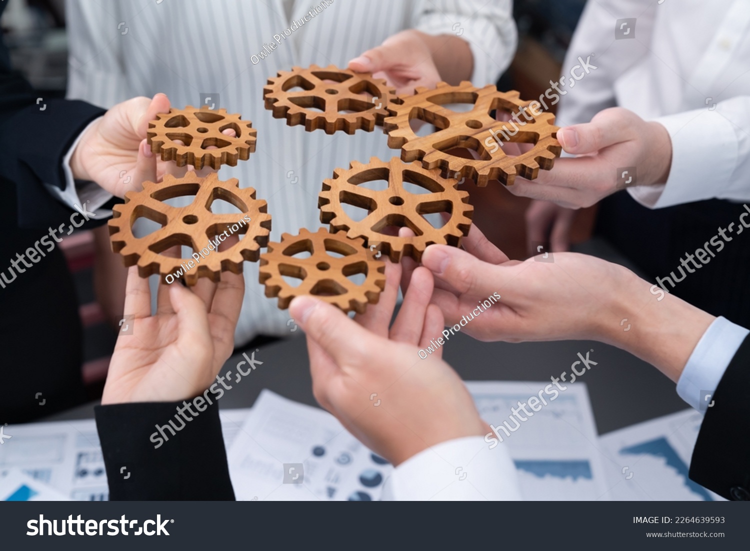 Closeup top view businesspeople hand holding gear and join together over meeting table with financial report papers. Cohesive group of office workers holding cog wheel as synergy harmony concept. #2264639593