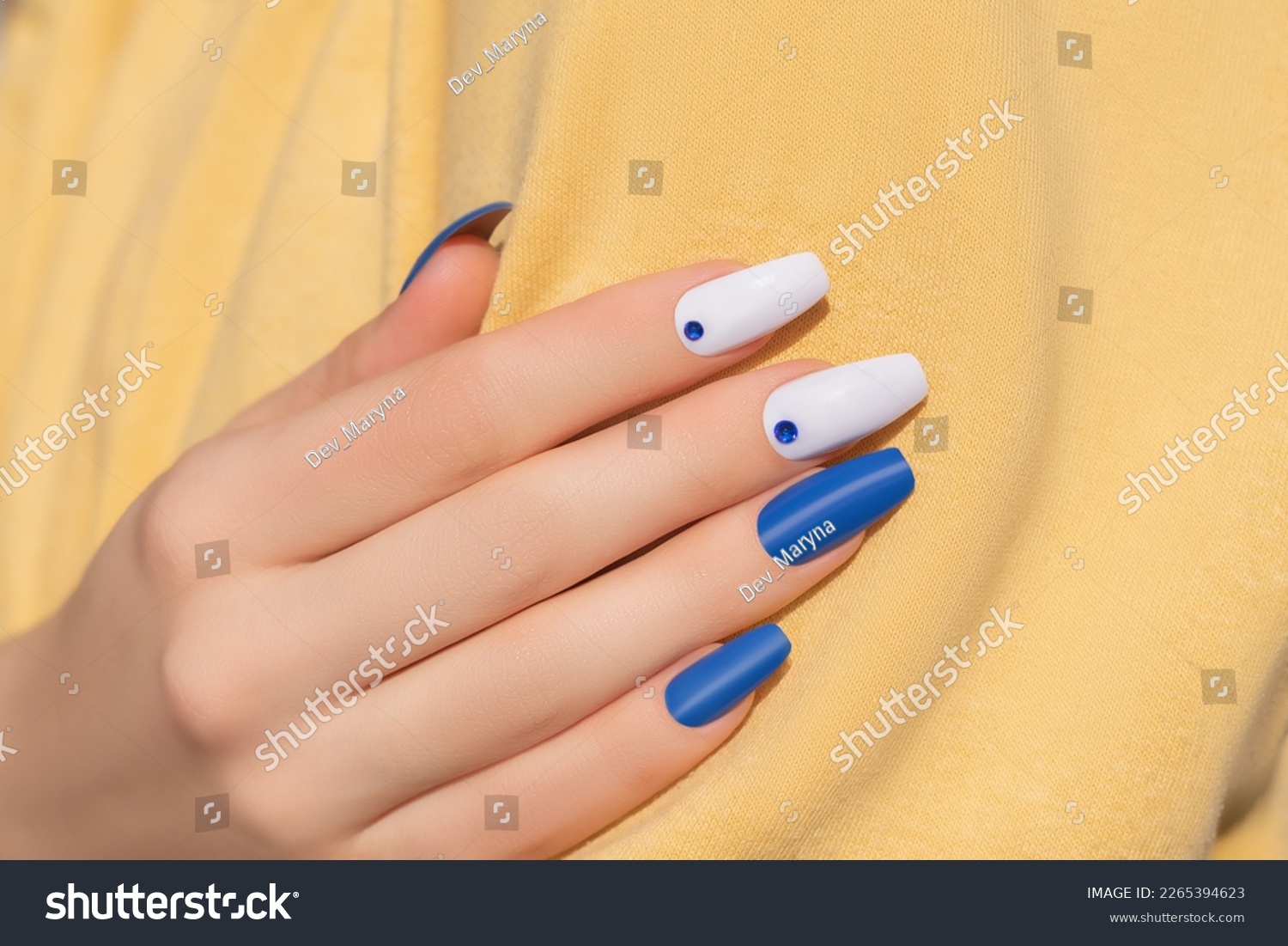 Female hand with rhinestones nail design. Glitter white and blue nail polish manicure with rhinestones nail art. Female model hand with perfect manicure and nail art on yellow fabric background. #2265394623
