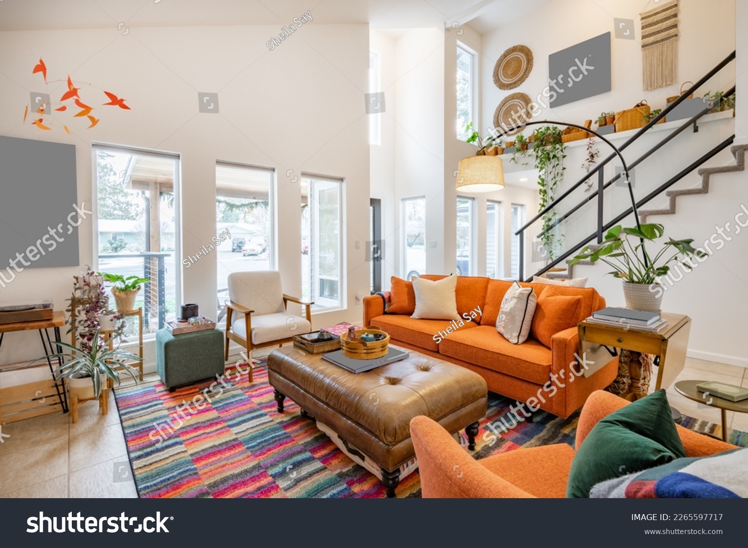 Bohemian style living room with orange sofa colored chairs books houseplants stair case and cluttered decor #2265597717