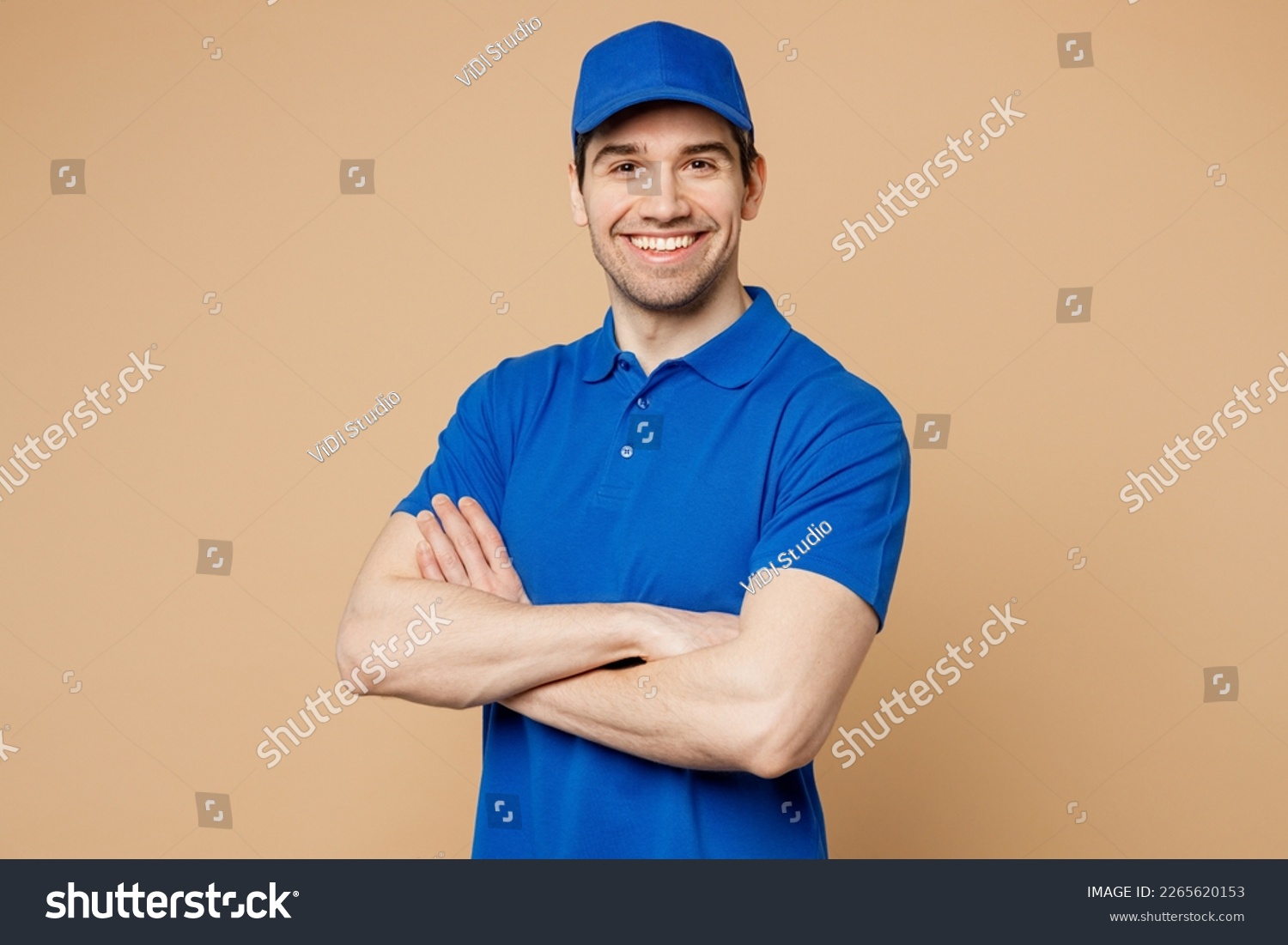 Professional delivery guy employee man wears blue cap t-shirt uniform workwear work as dealer courier look camera hold hands crossed folded isolated on plain light beige background. Service concept #2265620153