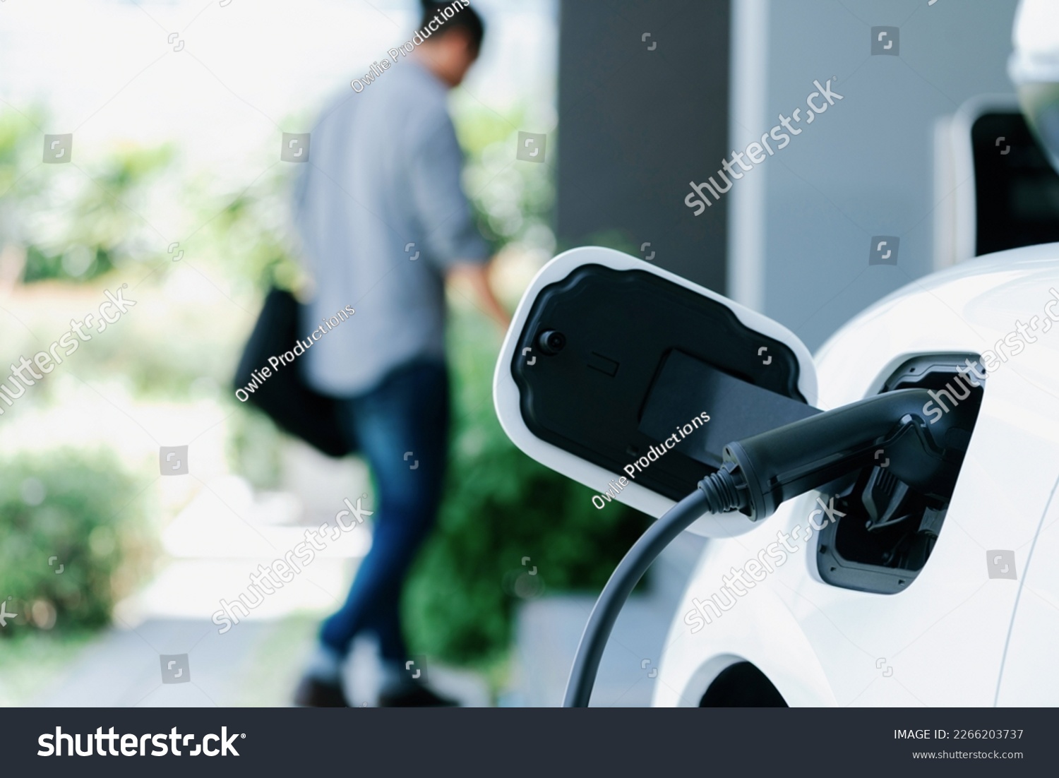 Focus electric car charging at home charging station with blurred progressive man walking in the background. Electric car using renewable clean for eco-friendly concept. #2266203737