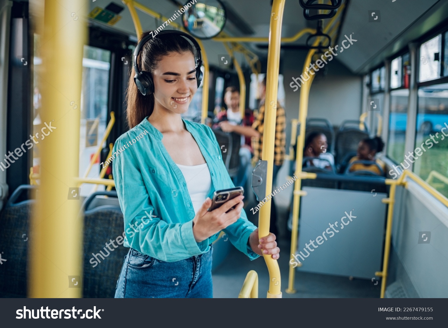 Young woman smiling while standing by herself on a bus and listening to music on a smartphone. Happy female passenger using headphones and a mobile phone in public transportation. Copy space. #2267479155