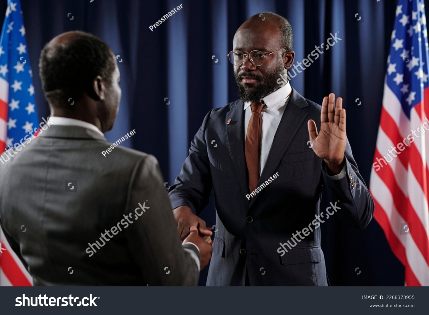 Serious young African American male president pronouncing oath of office with his right hand on Holy Bible while keeping open left one #2268373955