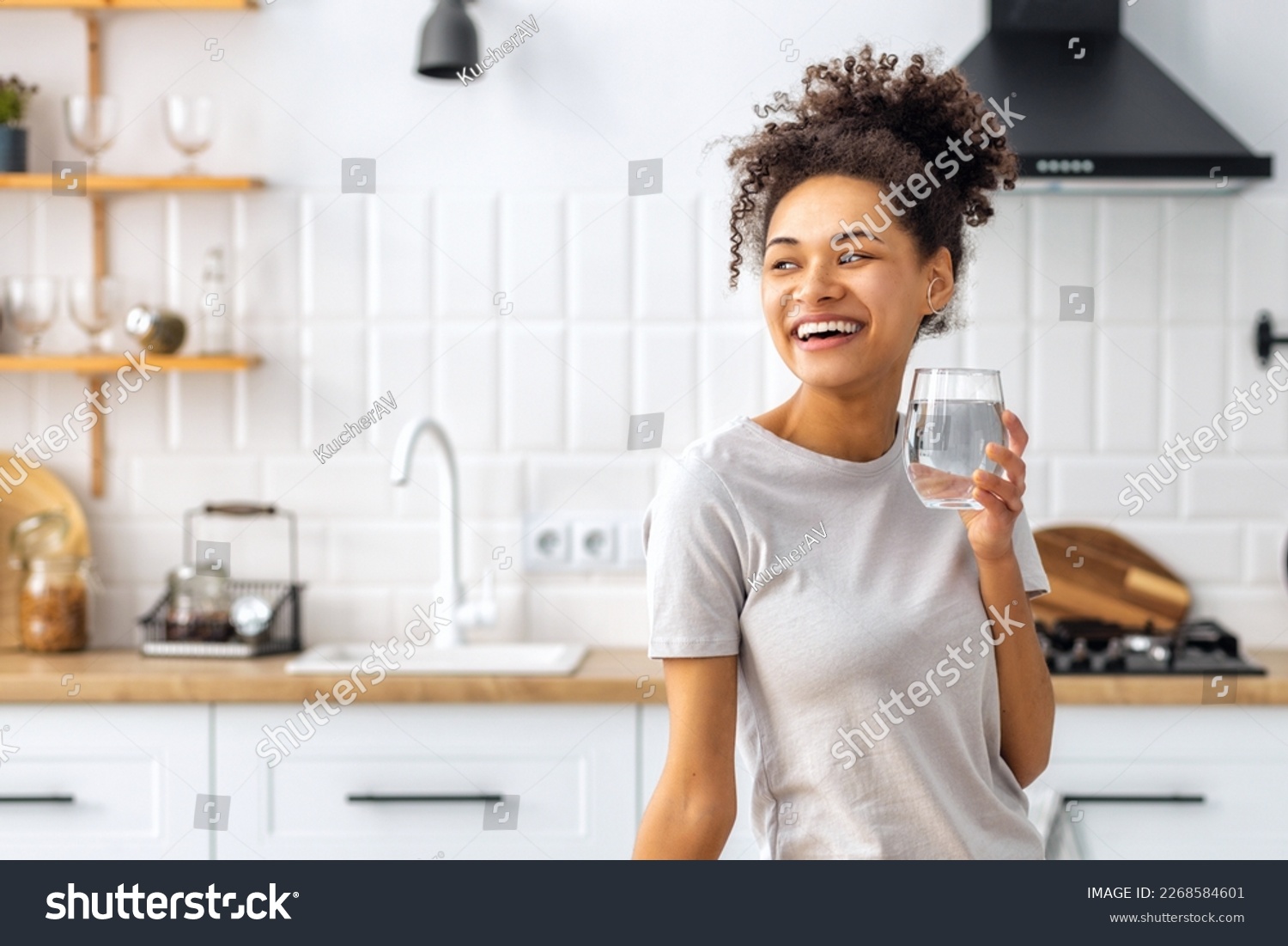 Portrait of a beautiful African American young woman drinking water standing in the kitchen at home looking away and smiling, wellness healthy food concept #2268584601