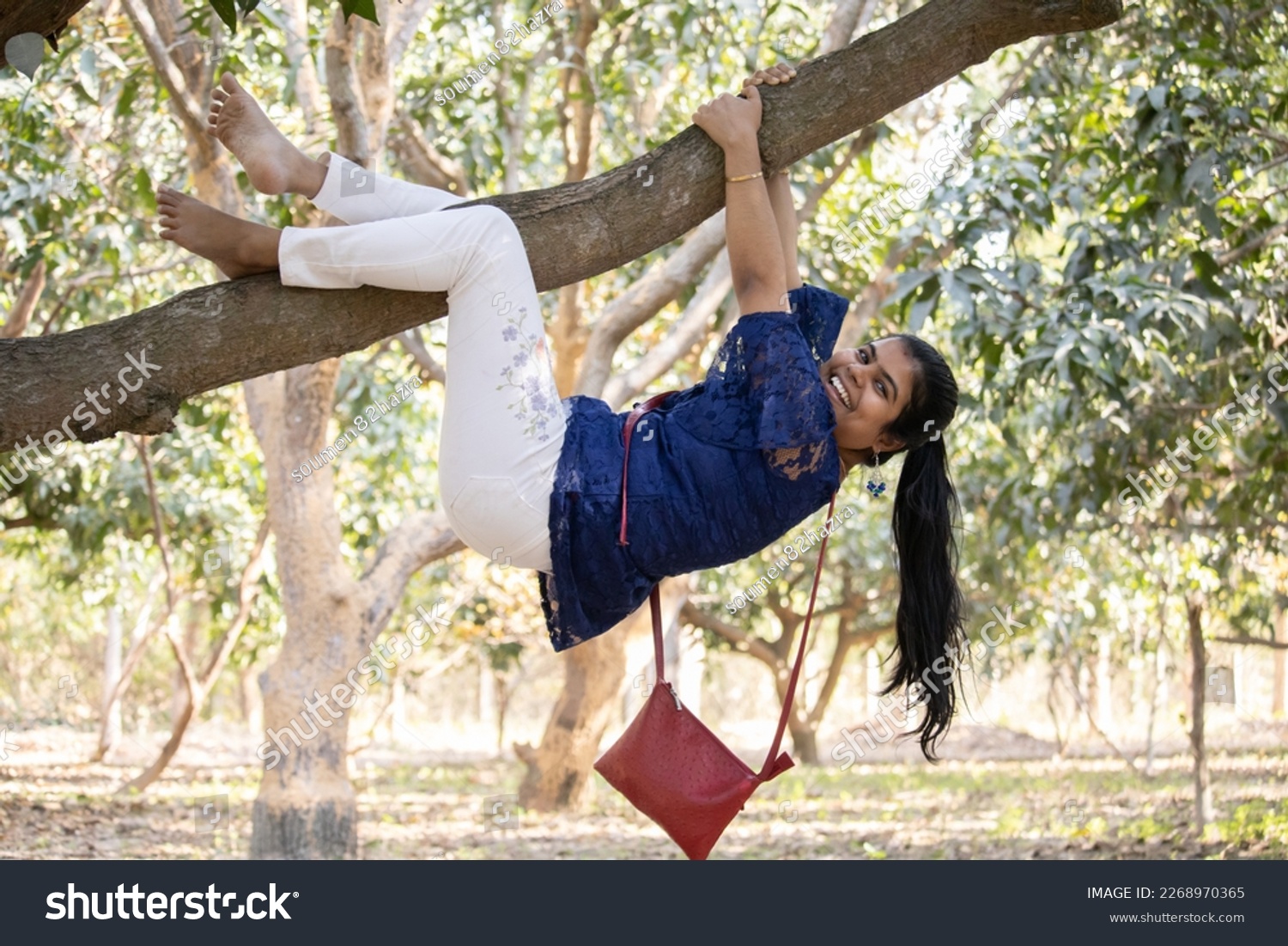 An indian young adult girl playfully climbing a tree in garden #2268970365