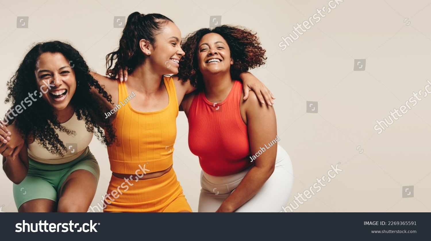 Group of three young, diverse female athletes celebrate their healthy and active lifestyle in a sports studio, smiling and laughing together while wearing sporty fitness clothes. #2269365591