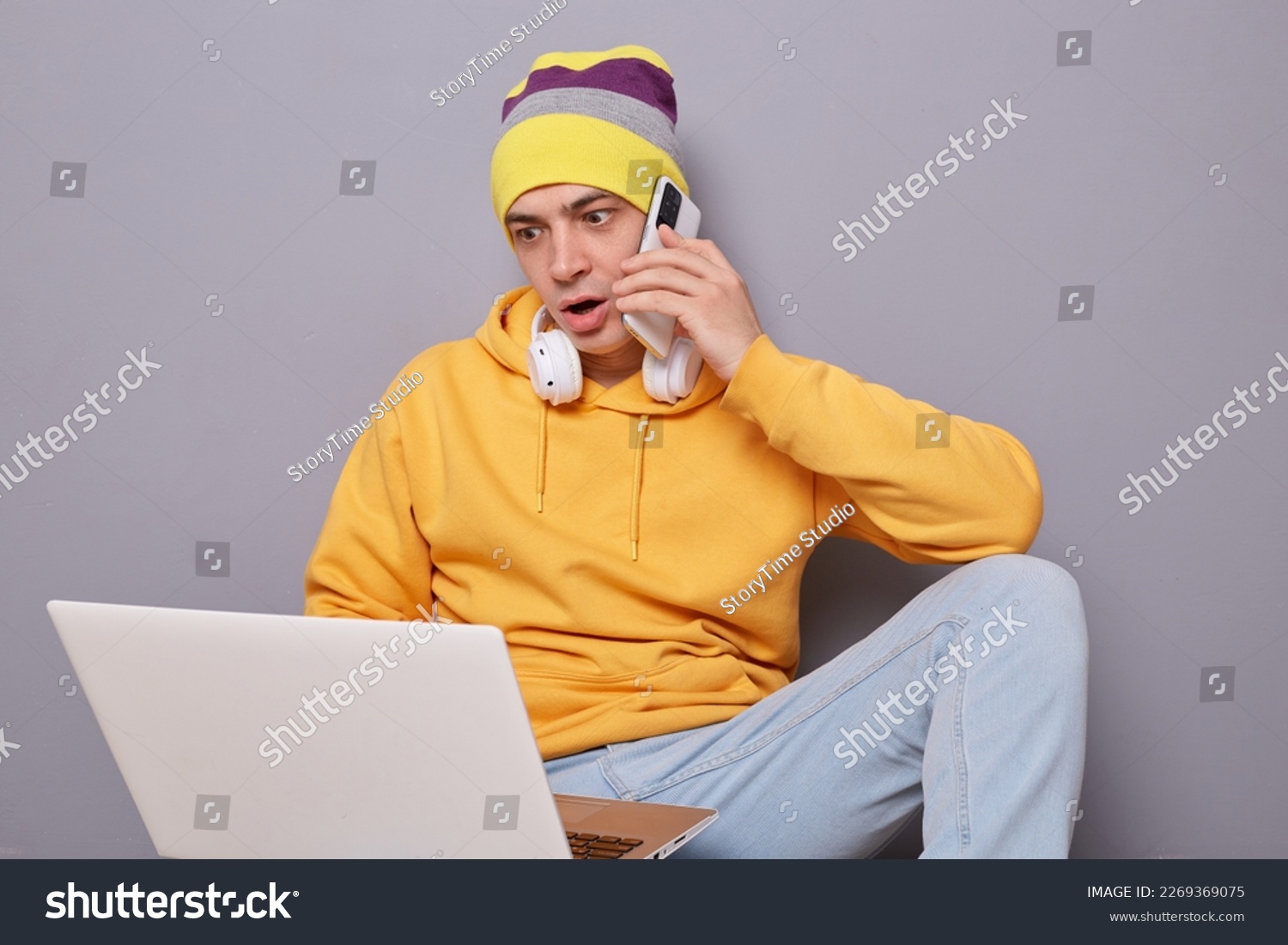Portrait of shocked man wearing beanie hat, yellow hoodie and jeans posing against gray wall, looking at laptop monitor with big eyes, talking on cell phone with somebody. #2269369075