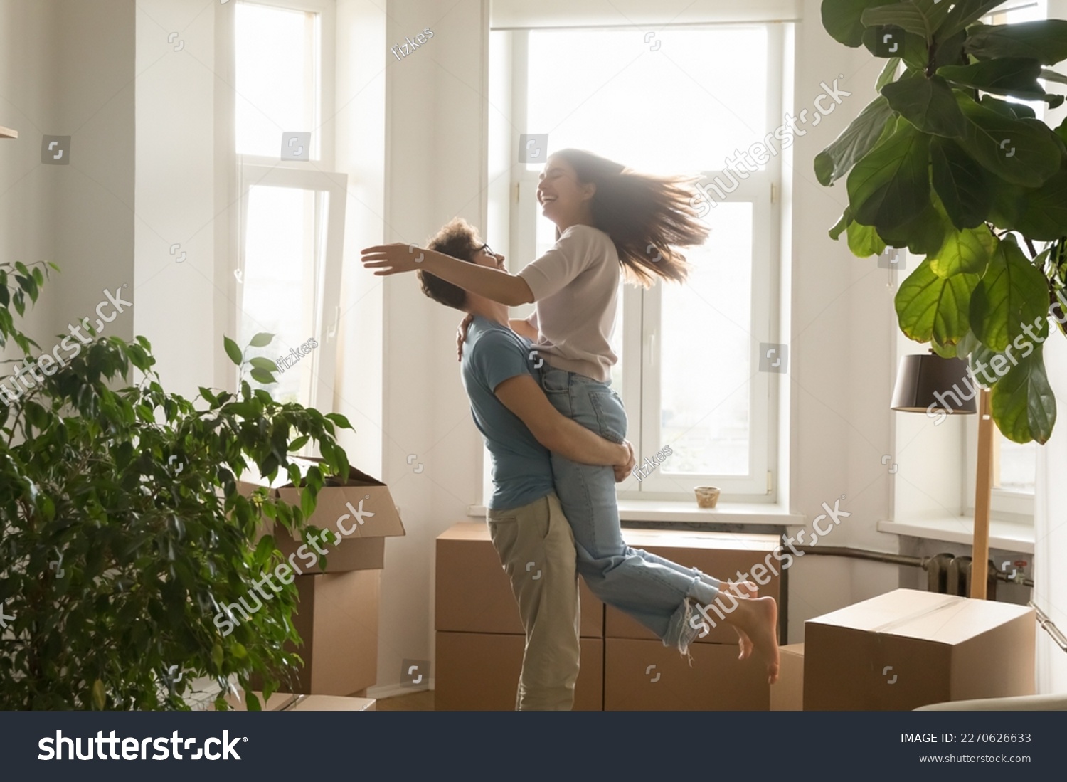 Happy excited young couple celebrating relocation, buying flat, apartment, moving in together, dancing, having fun, laughing. Guy holding girlfriend in arms, spinning at cardboard boxes #2270626633