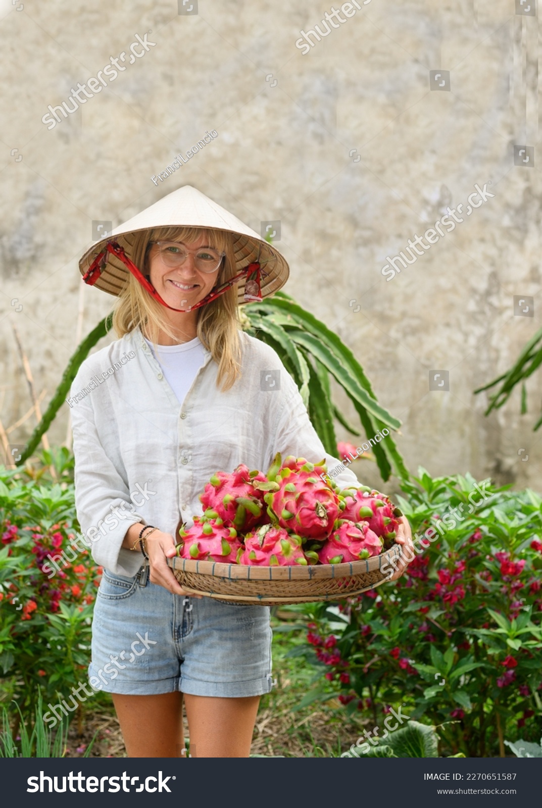 Positive adult female in white shirt and conical hat standing in garden and carrying wicker basket with dragon fruits #2270651587