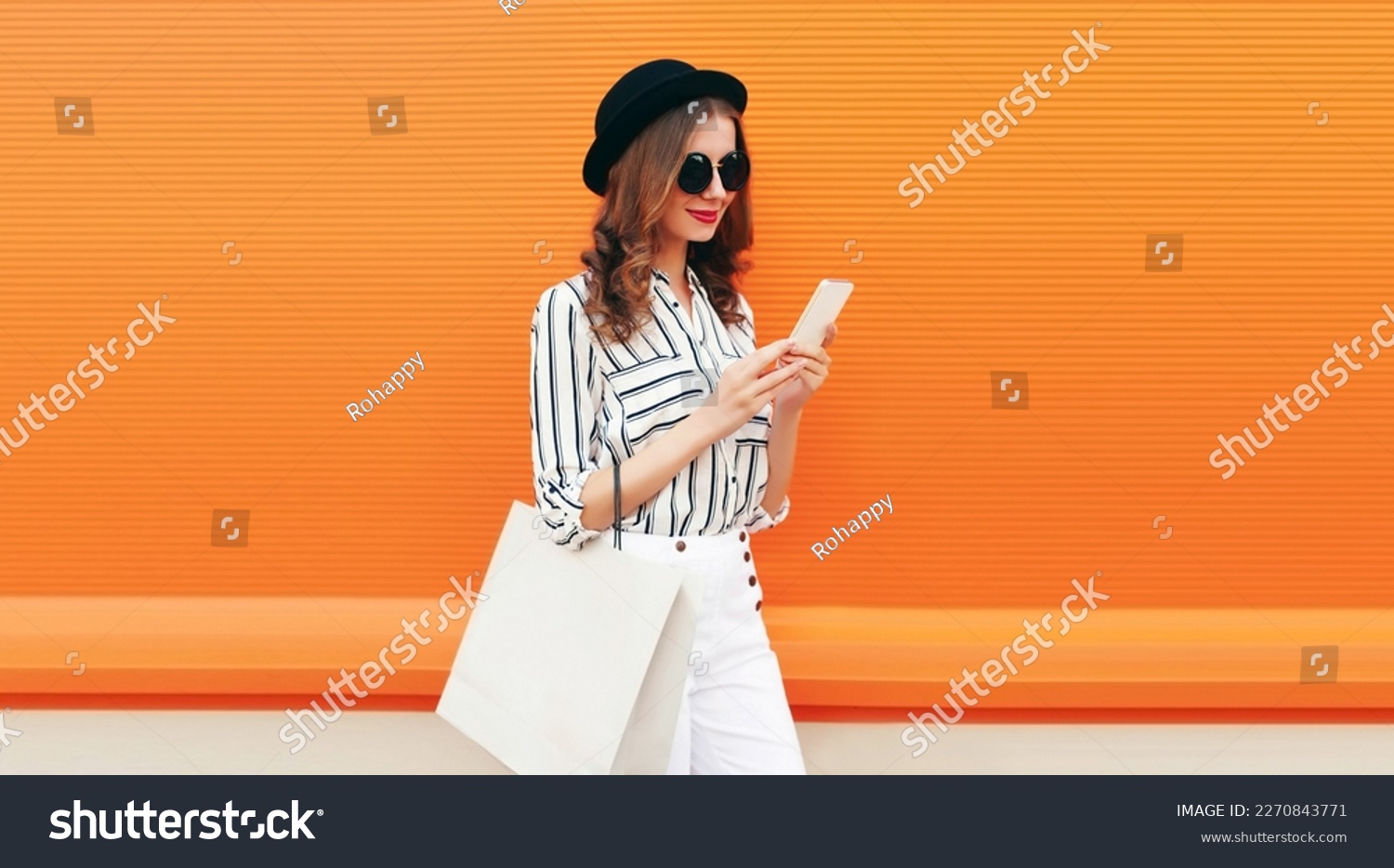 Portrait of beautiful young woman looking at smartphone with shopping bag wearing white striped shirt, black round hat on orange background #2270843771