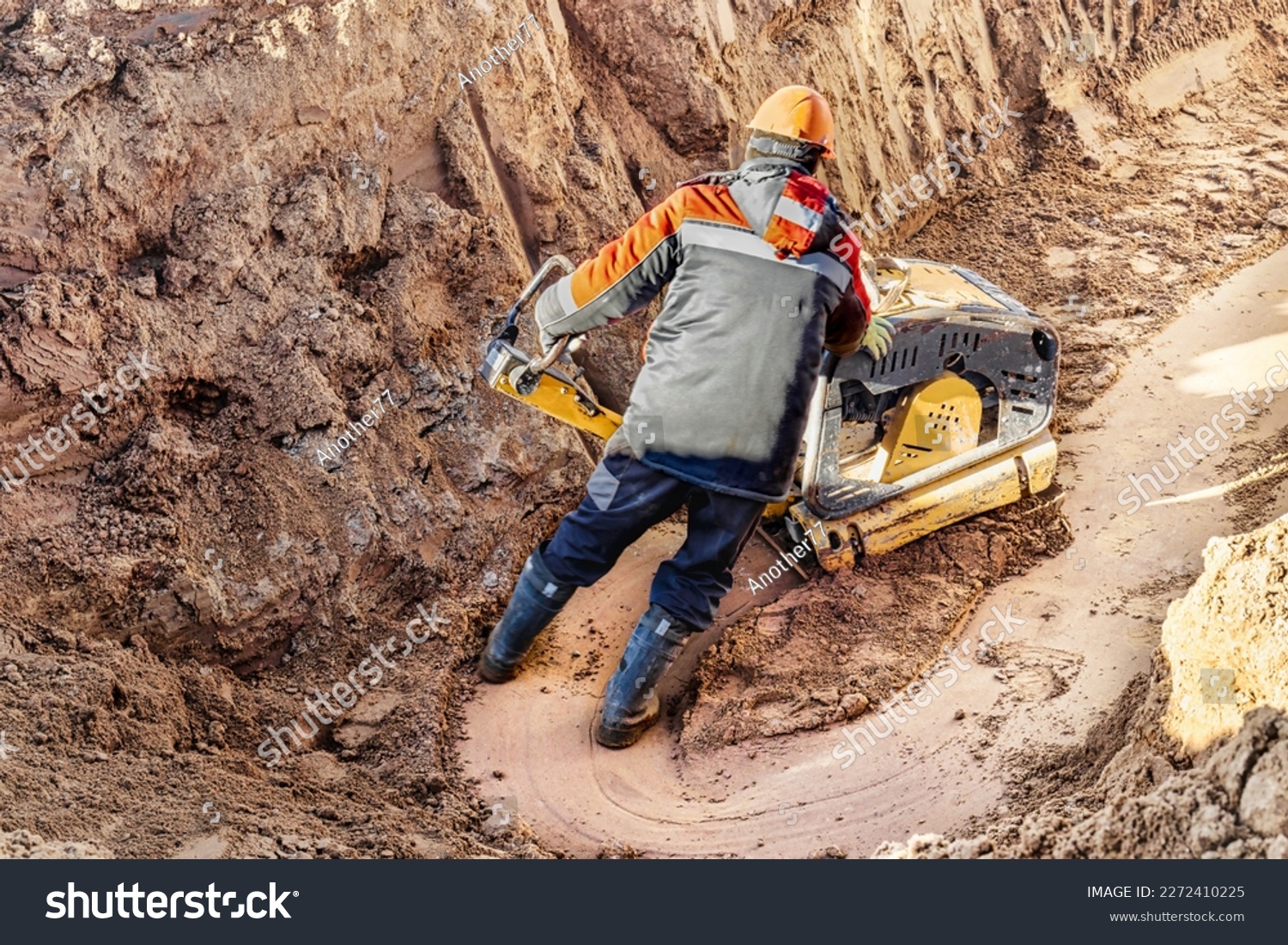 A worker compacts soil or sand with a vibrating plate in a trench at a construction site close-up. Vibratory soil compaction for laying underground utilities #2272410225