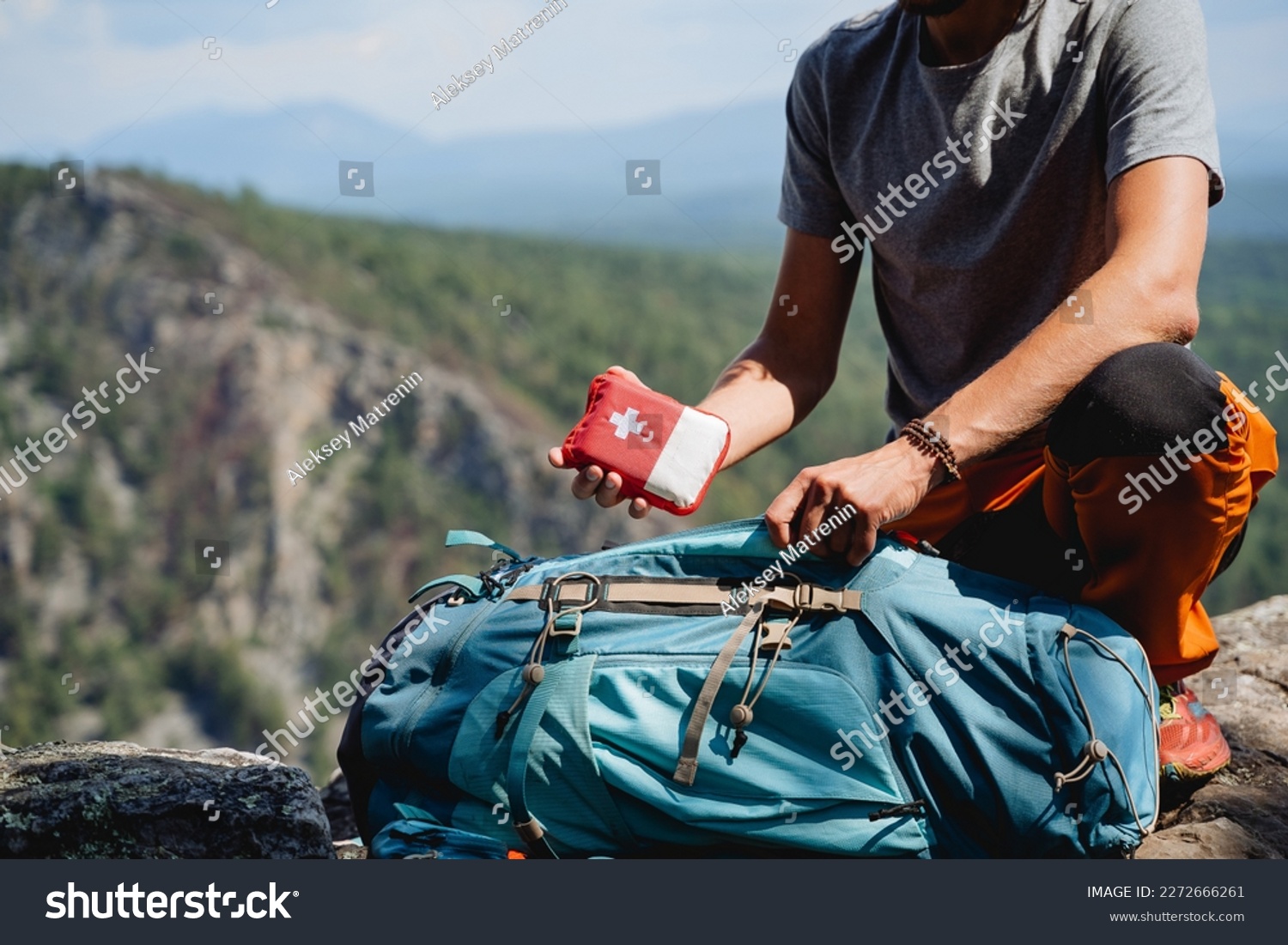 Take a first-aid kit in a backpack on a trip, a hand holds a first aid kit against the background of mountains, equipment in an extreme hike, a white cross #2272666261