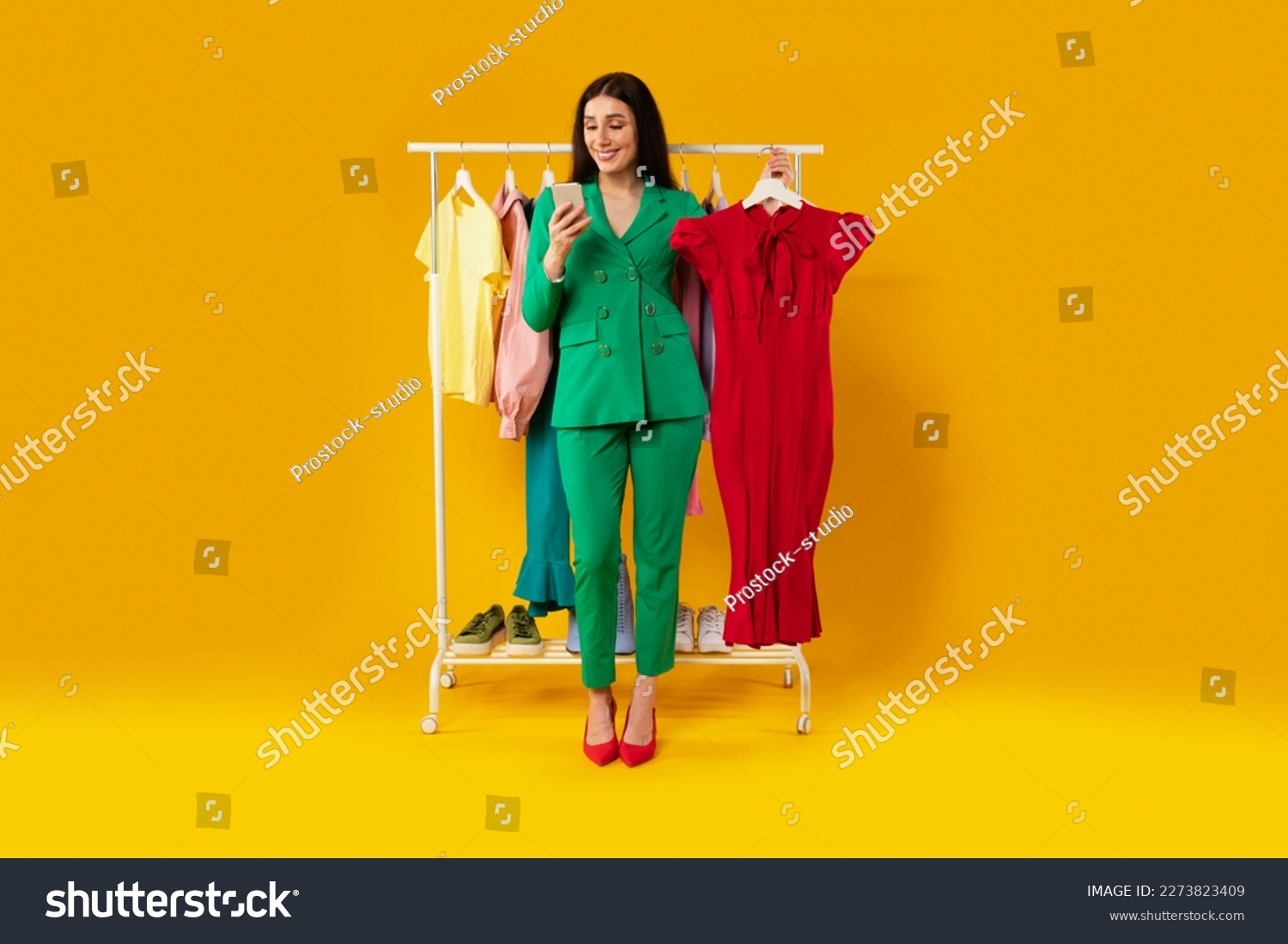 App for online shopping. Young european lady using cellphone and holding dress, ordering clothes online in mobile app over yellow studio background, full length #2273823409