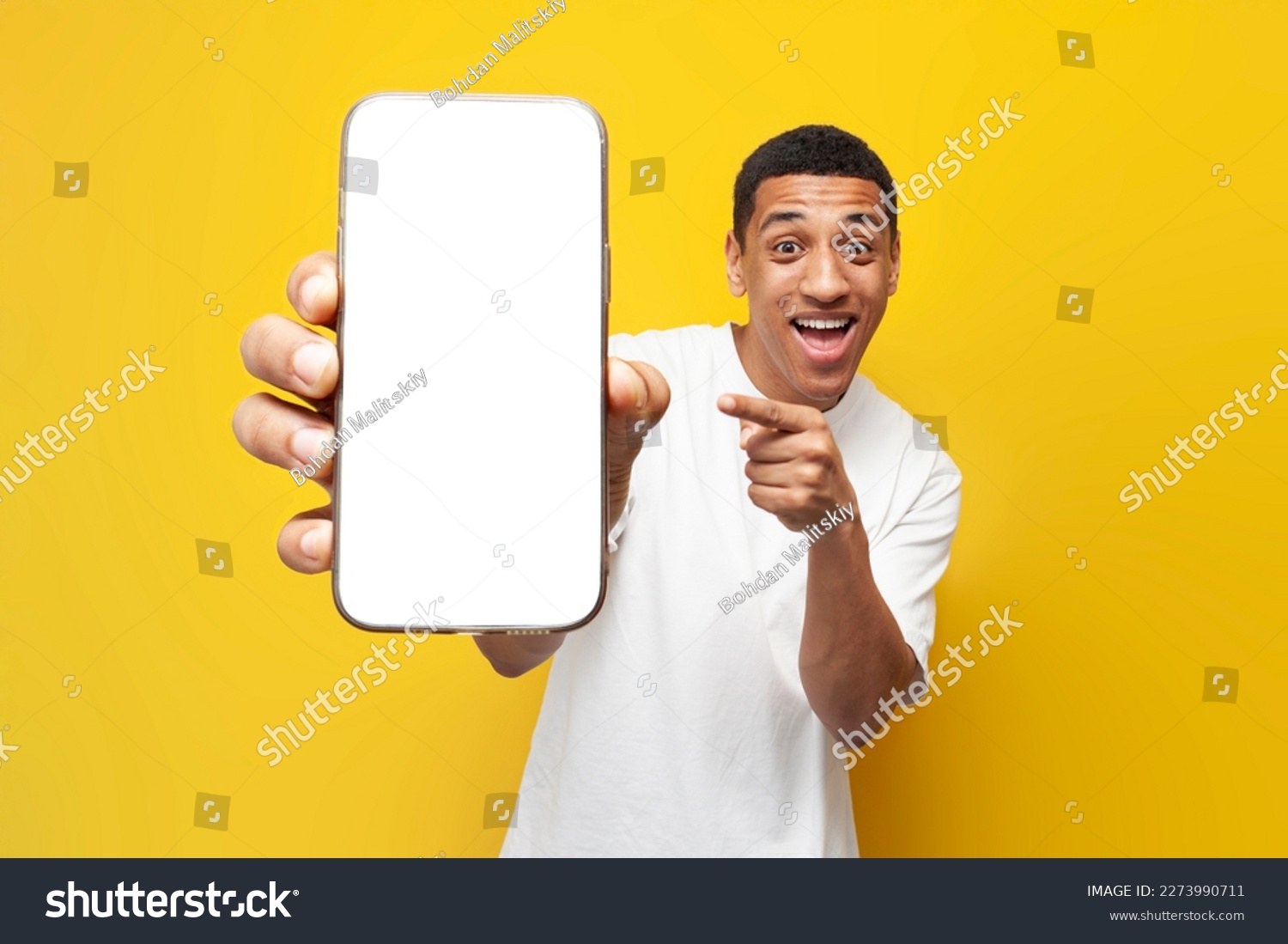 young african american guy in white t-shirt shows blank screen of smartphone on yellow isolated background, man holds phone and advertises screen for mock-up #2273990711