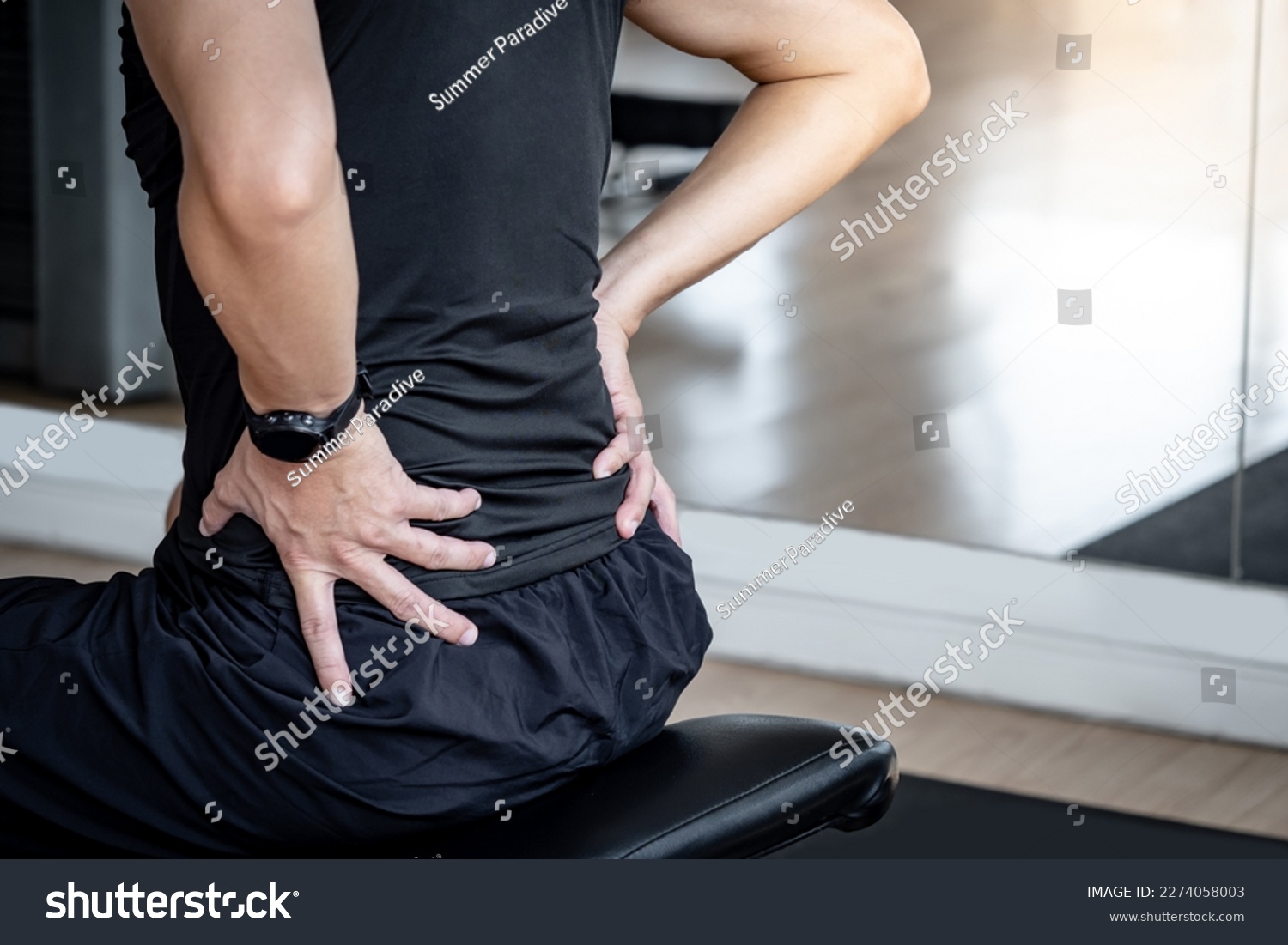 Sport man feeling lower back pain or spine pain while sitting on workout bench in fitness gym. Male athlete suffering from sport injuries symptoms. #2274058003