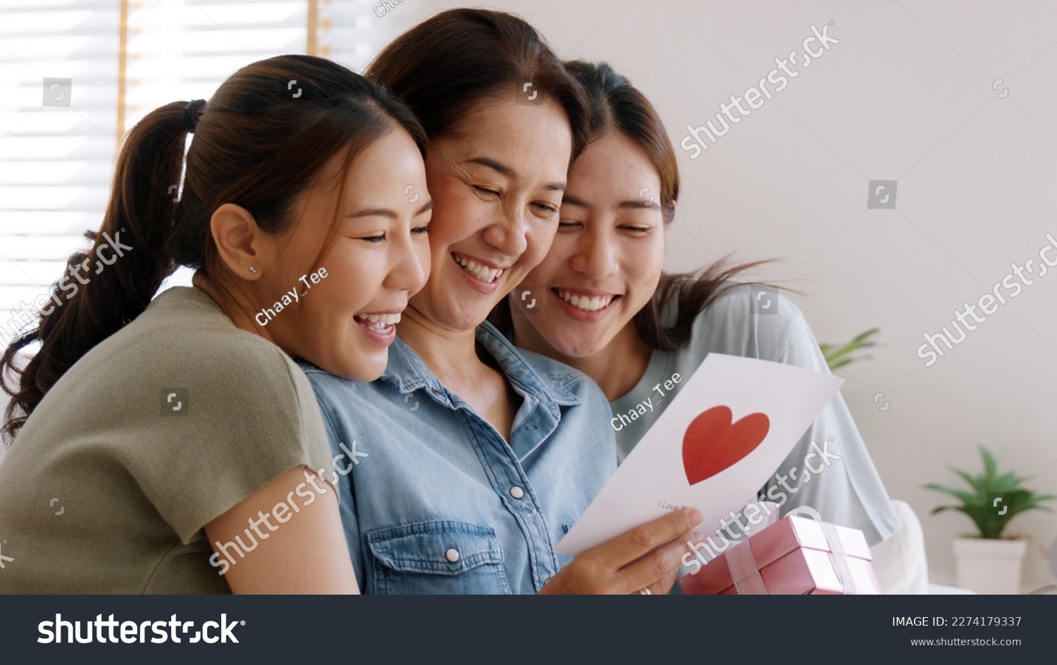 Mother day two grown up child cuddle hug give flower gift box red heart card to mature mum. Love kiss care mom asia middle age adult three people sitting at home sofa happy smile enjoy family time. #2274179337