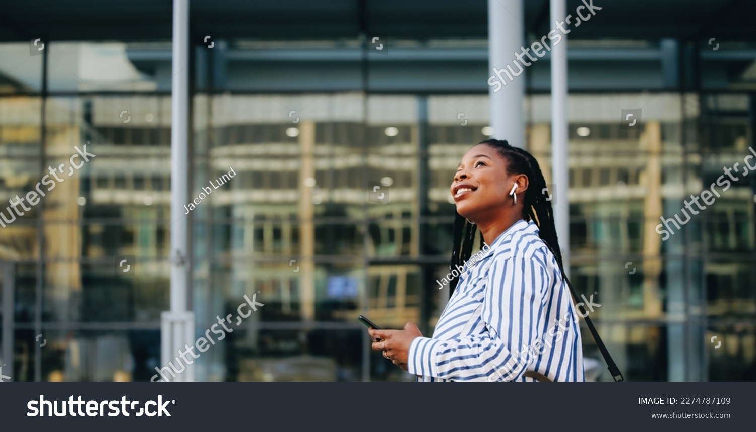 Sideview of a young businesswoman looking away and holding a smartphone while commuting in the city. Cheerful young business woman listening to music on her way to the office in the morning. #2274787109