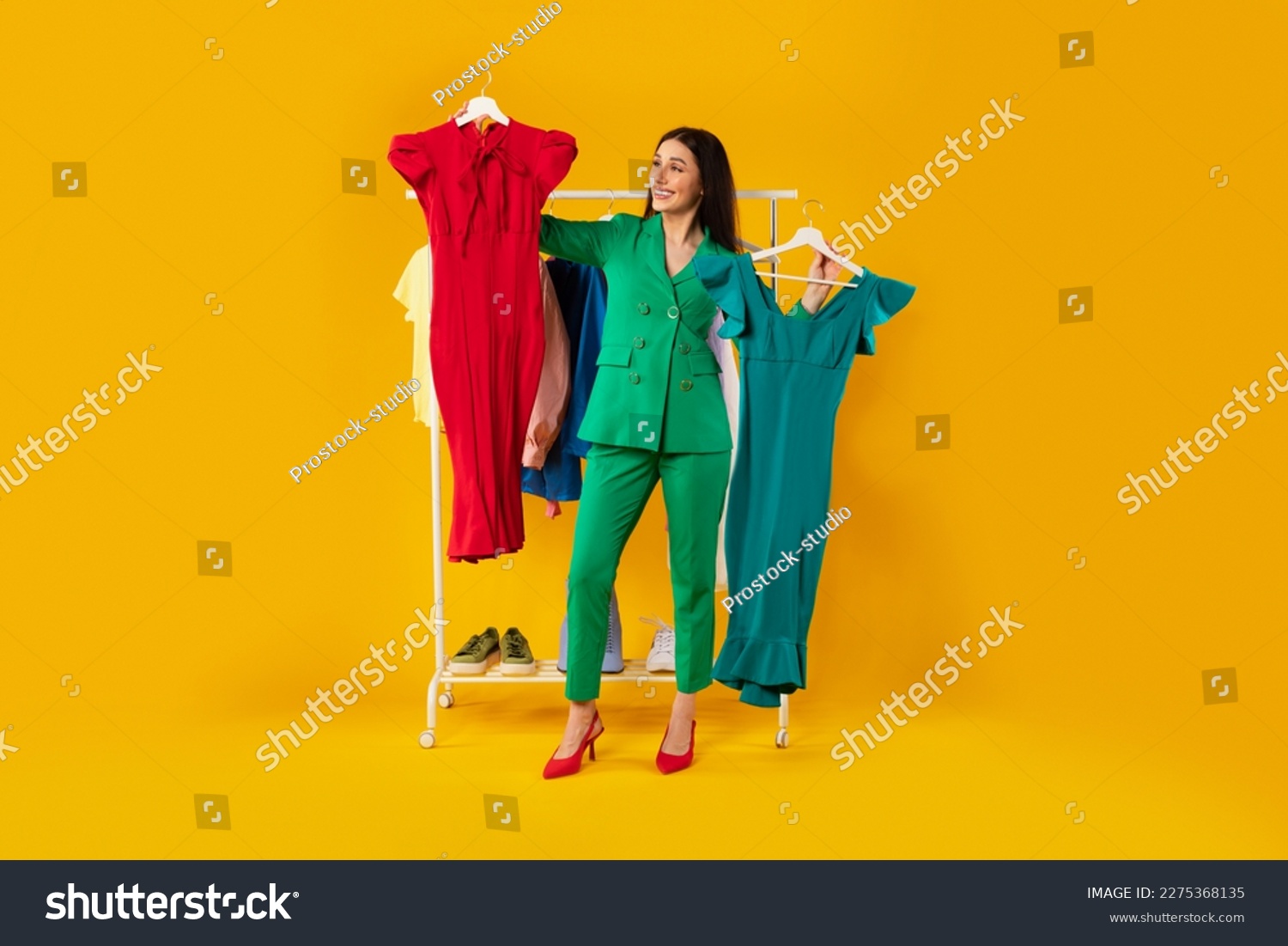 Shopaholic. Happy elegant woman choosing dresses holding two hangers, standing near clothing rail with trendy clothes over yellow background. Female fashion choice #2275368135