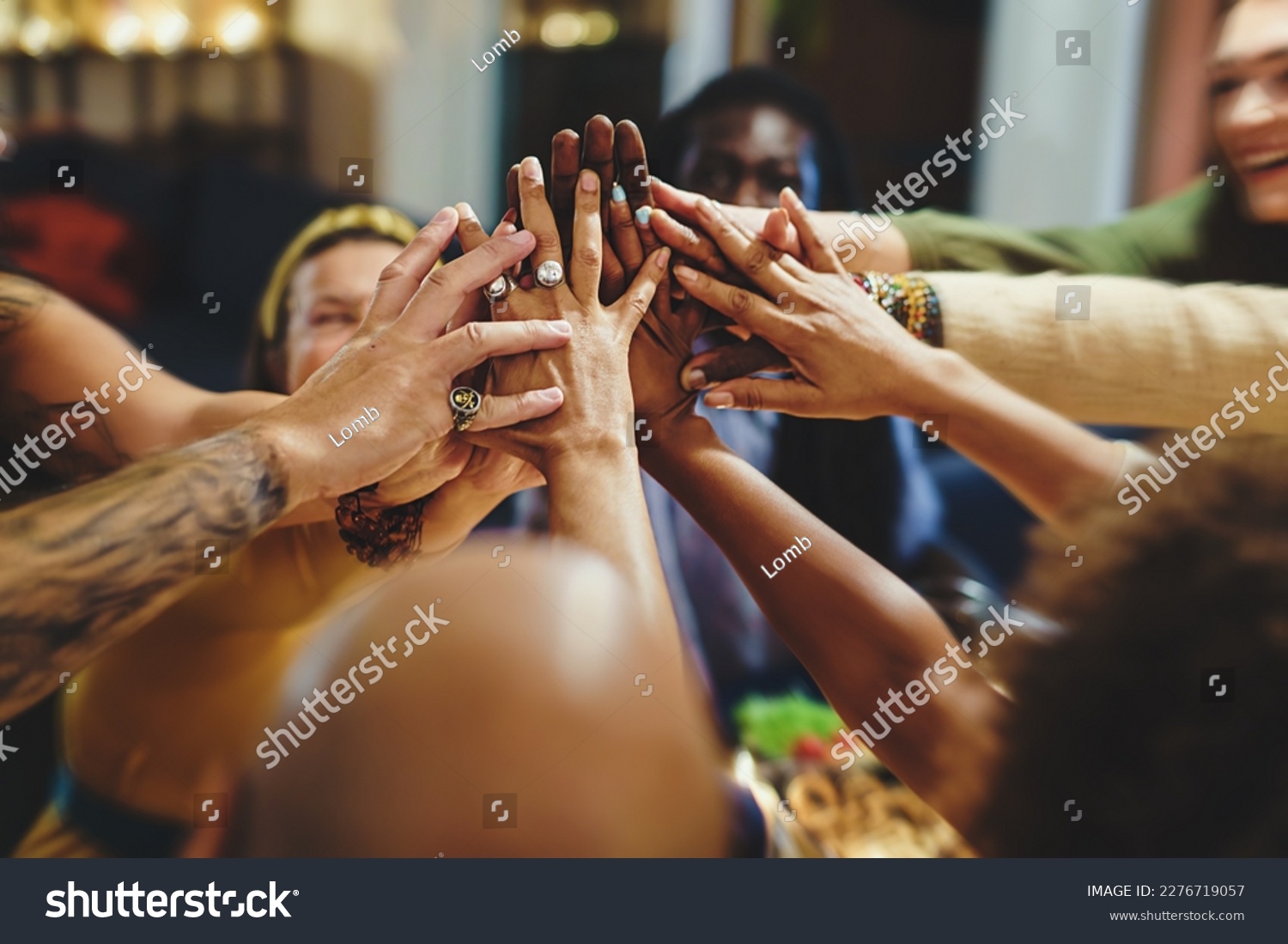 A diverse group of friends, in their forties, with various ethnicities and tattoos, gather around a table for dinner. They enthusiastically join hands in a high-five, blurred faces and focus on hands #2276719057