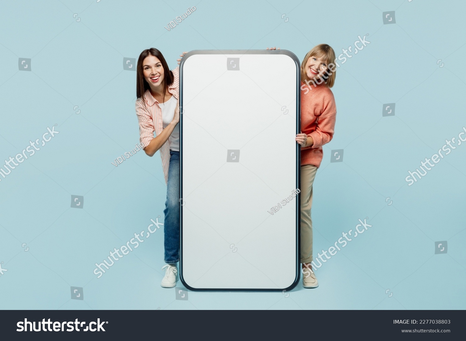 Full body elder parent mom with young adult daughter two women together in casual clothes big huge blank screen area mobile cell phone look camera isolated on plain blue background Family day concept #2277038803