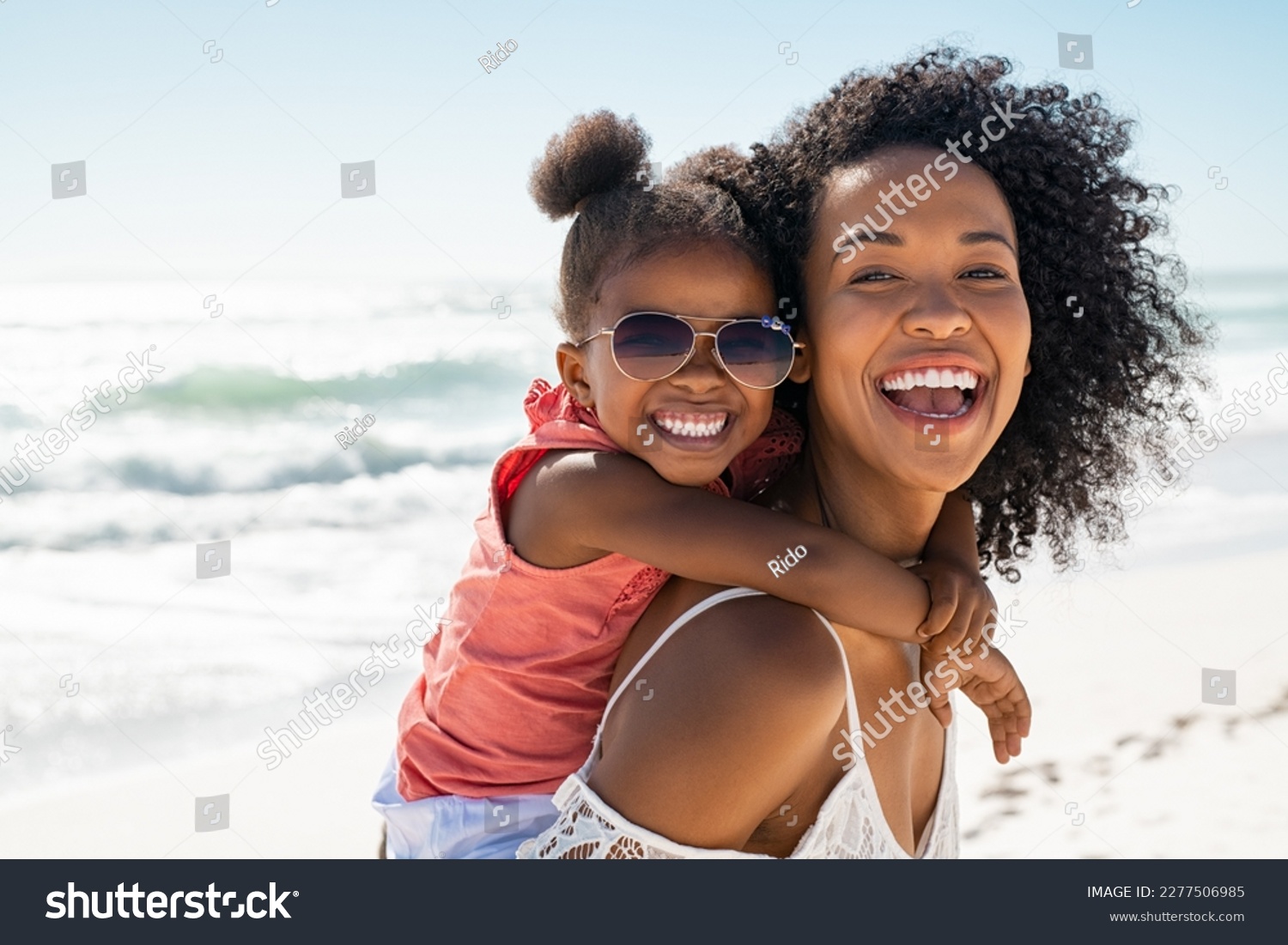 Smiling black mother and beautiful daughter wearing sunglasses having fun on the beach. Portrait of happy african american woman giving a piggyback ride to her cute little girl wearing shades. #2277506985