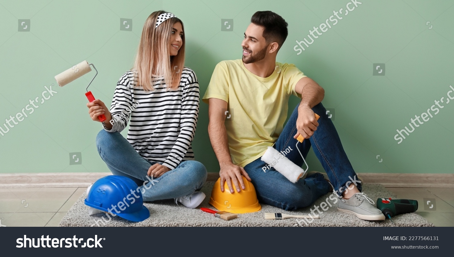 Happy young couple with paint rollers and hardhats sitting on floor in their new house #2277566131
