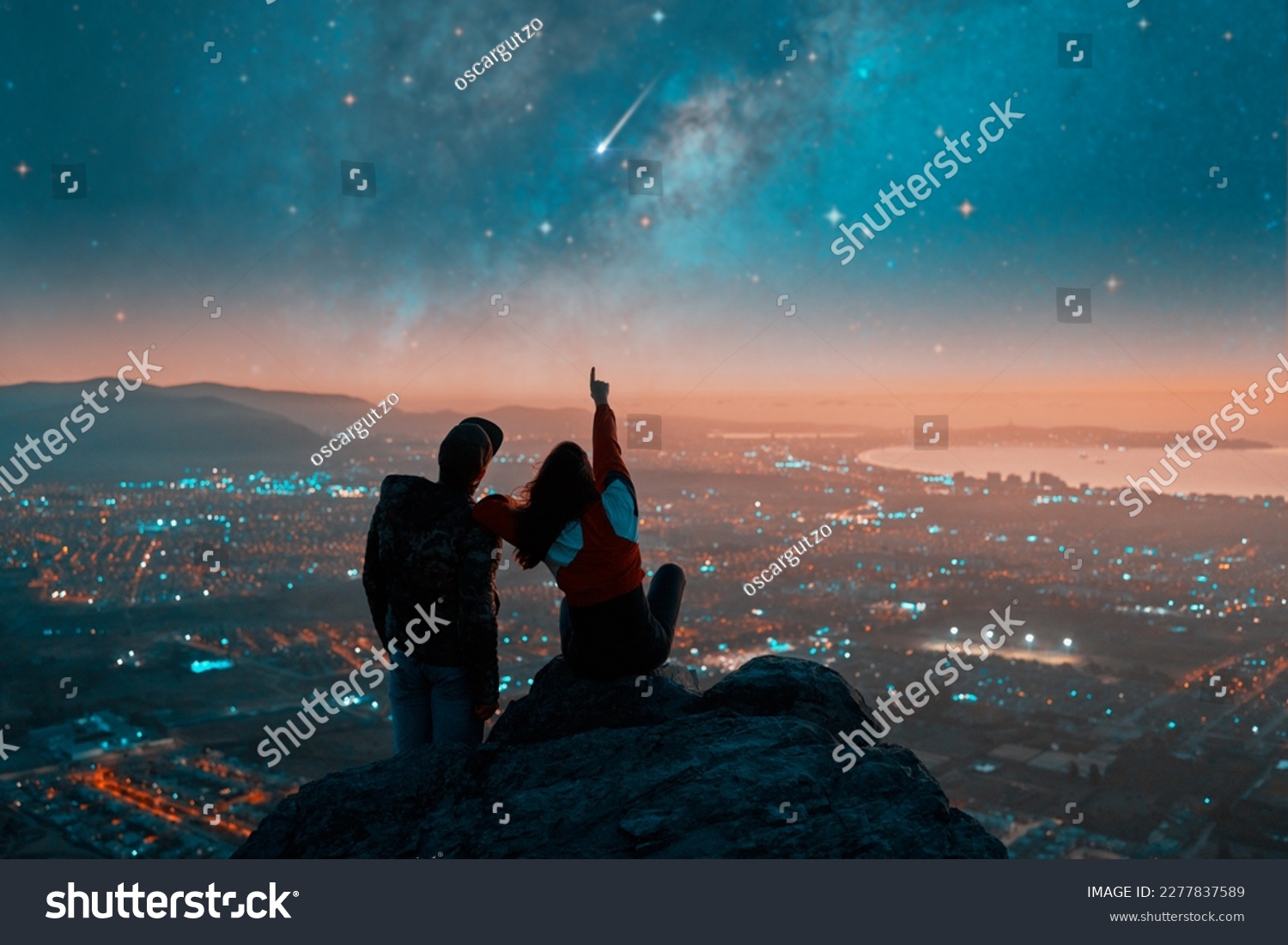silhouettes of a couple sitting on the top of the mountain looking and pointing out at shooting star and milky way over the city lights on the horizon	 #2277837589