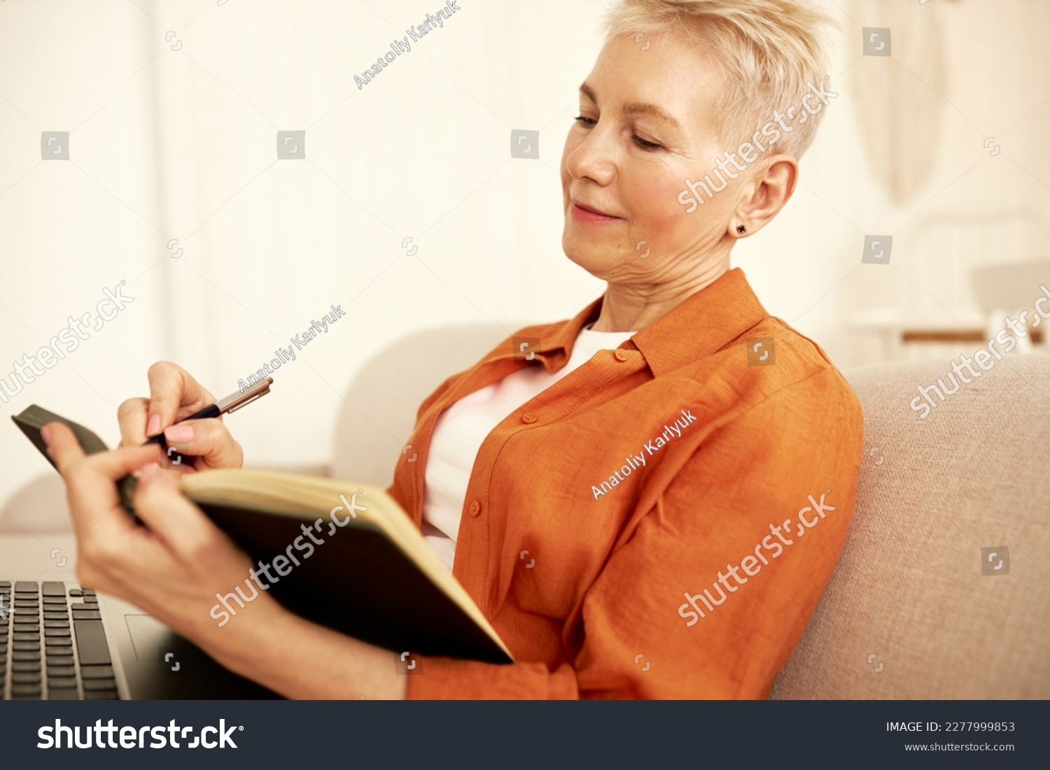 Side view of stylish aged female novelist sitting on couch with laptop on knees noting down thoughts and ideas for new book. Mature woman writing in her diary while sitting on sofa #2277999853