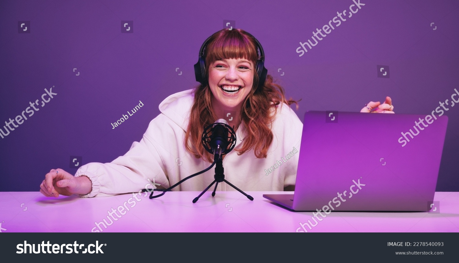 Happy young woman laughing while recording a live radio show in a studio. Cheerful young woman hosting an audio broadcast in neon purple light. Young woman creating content for her internet podcast. #2278540093