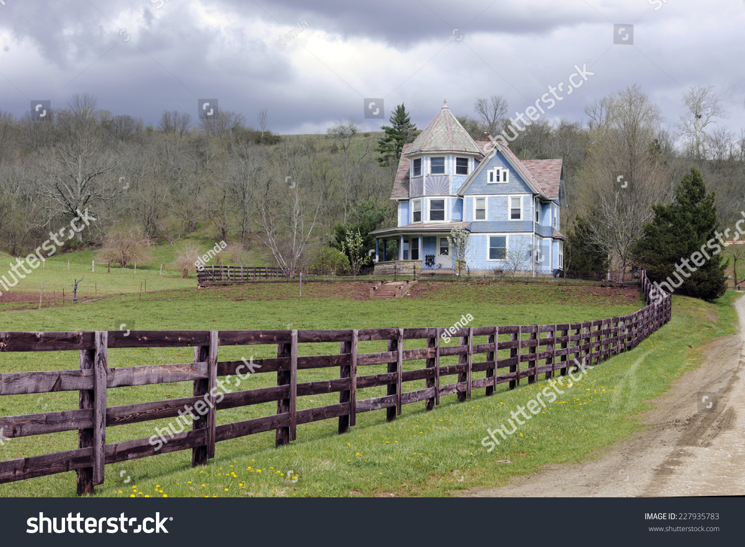 A rustic fence and dirt road by an old Victorian home under an overcast sky in the early spring.   #227935783