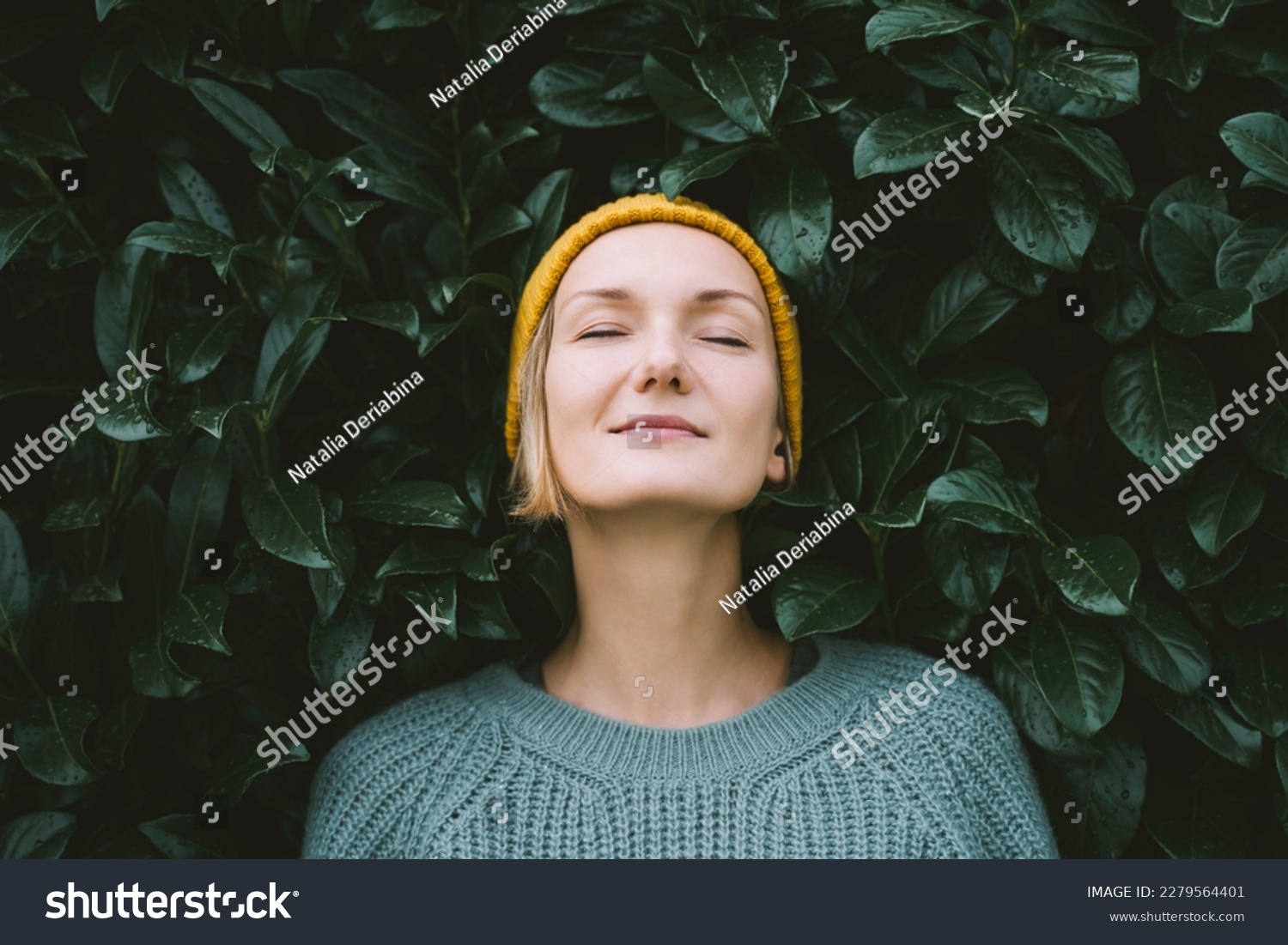 Portrait of relaxed and confident woman with eyes closed on background of green leaves wall. Thoughtful person in front of green hedge. Joy, zen and balance people. Stability through mental health #2279564401