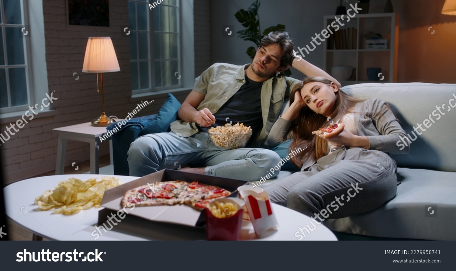 Caucasian extremely frustrated couple sitting in front of tv watching shows and eating pizza in absolute boredom. Student couple tired after a long day  #2279958741