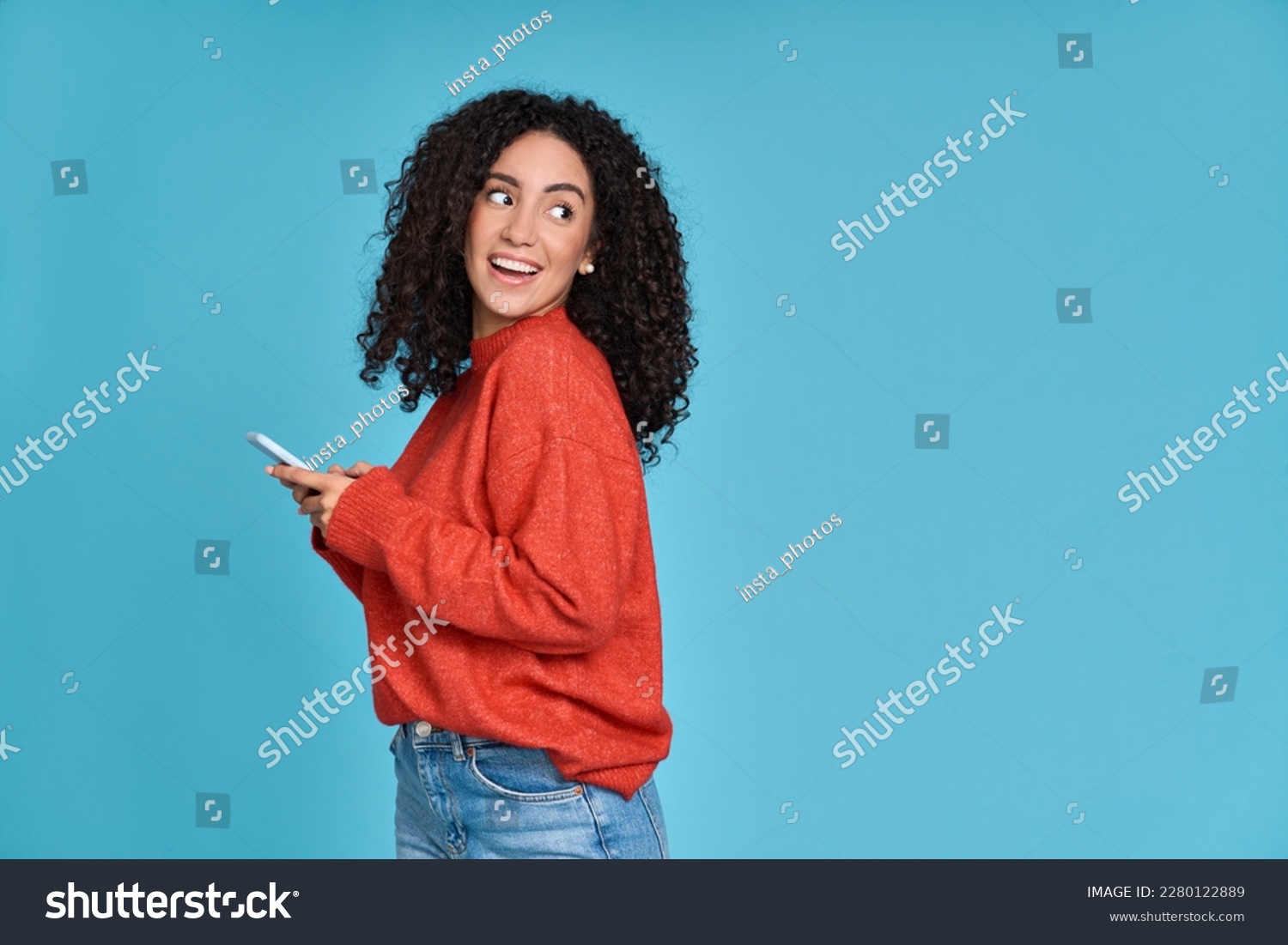 Young curious smiling happy pretty latin woman holding mobile phone, doing online shopping on cell, using apps on cellphone looking aside at copy space standing isolated on blue background. #2280122889