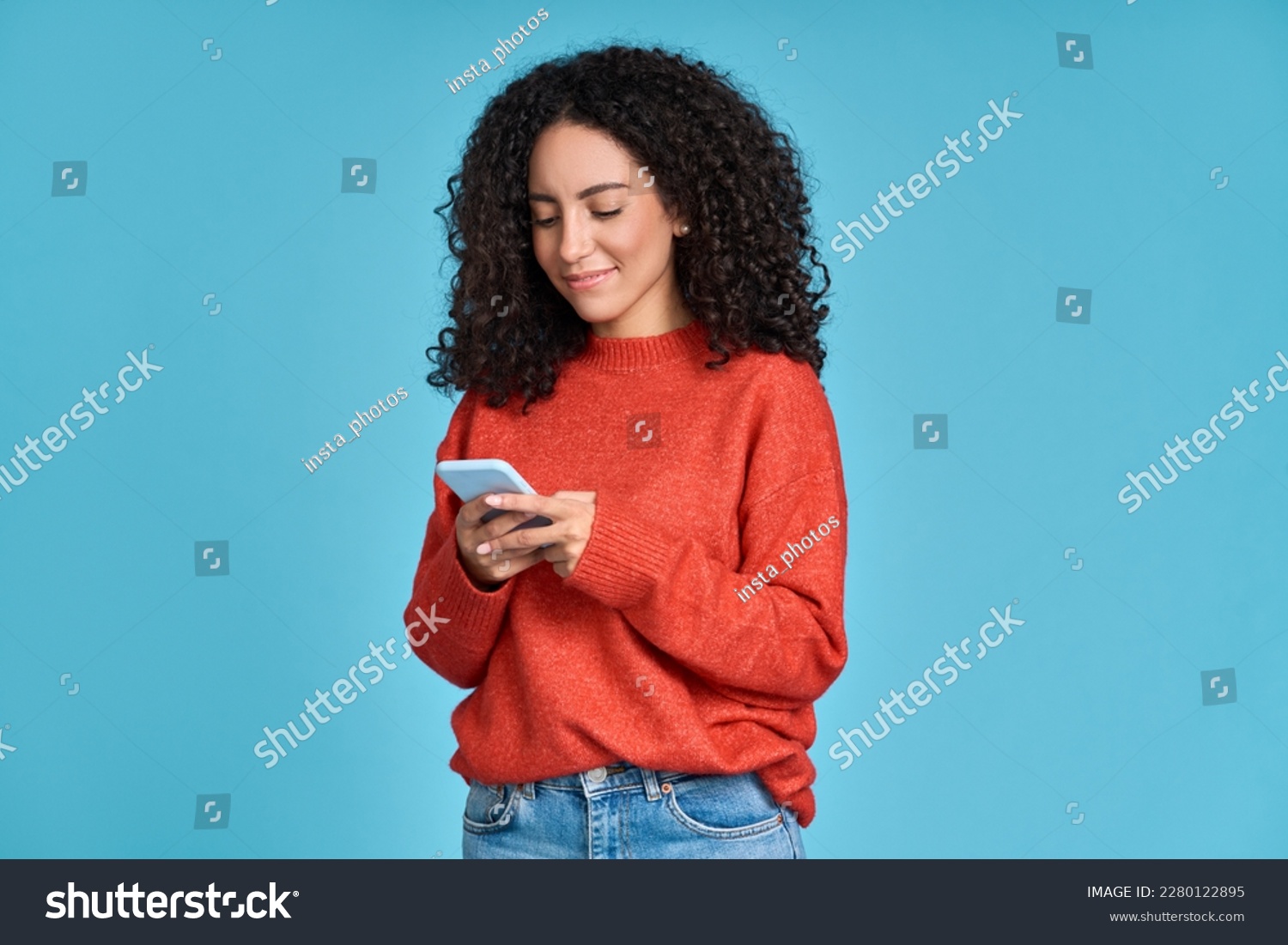 Young adult smiling happy pretty latin woman holding mobile phone looking at smartphone, typing message doing ecommerce shopping on cell, using trendy apps on cellphone isolated on blue background. #2280122895