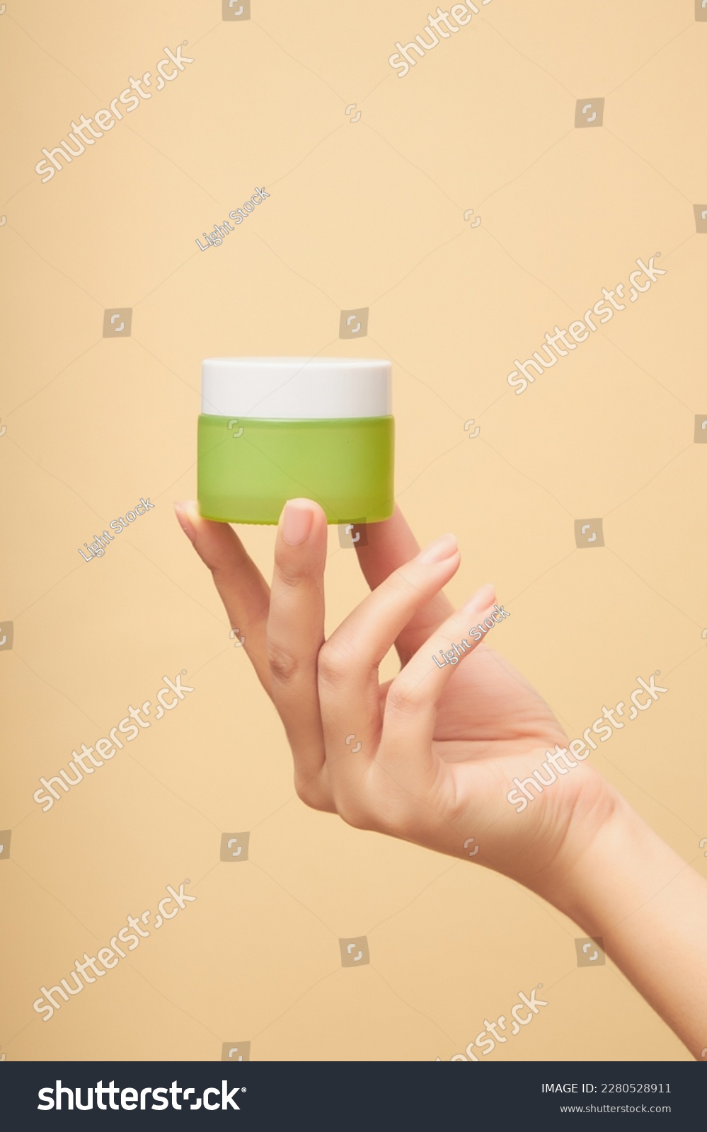 On the palm of the woman's hand is a moisturizing cream made of green glass on a beige background. Unlabeled bottle mockup of anti-aging masks, exfoliating cosmetics. Skin care concept #2280528911
