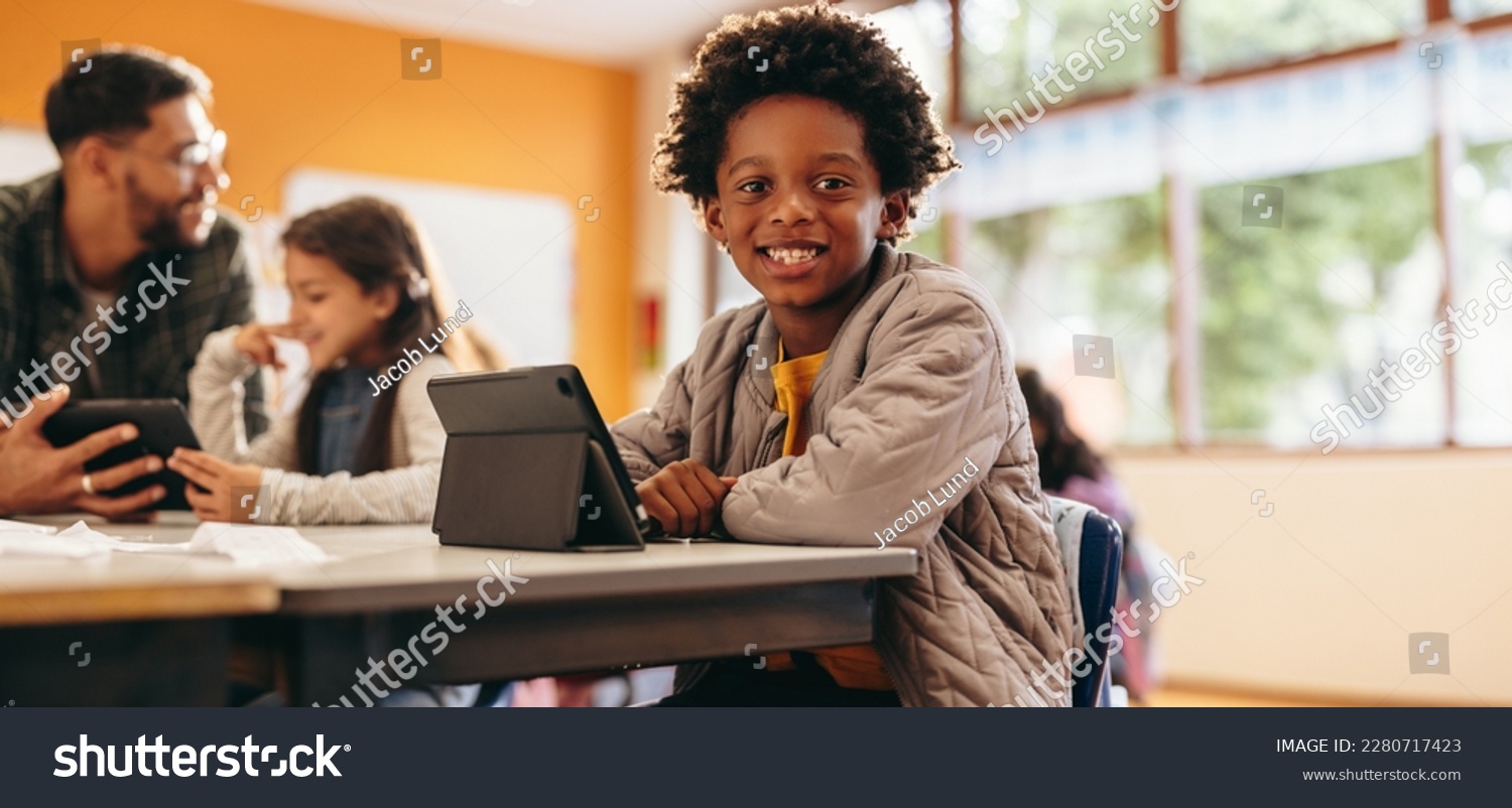 Boy smiling at the camera while sitting with a digital tablet in a classroom. Primary school kid reads an ebook at school. Young child developing his reading skills using modern technology. #2280717423