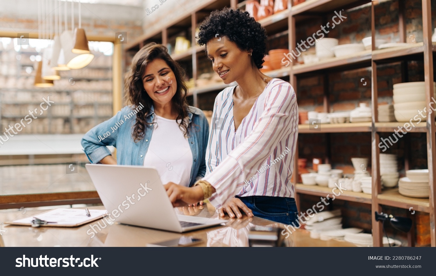 Successful female ceramists using a laptop while working together. Two female entrepreneurs managing online orders in their store. Happy young businesswomen running a creative small business together. #2280786247