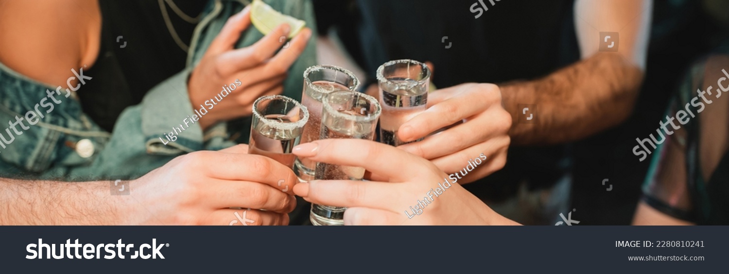 Cropped view of interracial friends holding glasses of tequila in bar, banner #2280810241