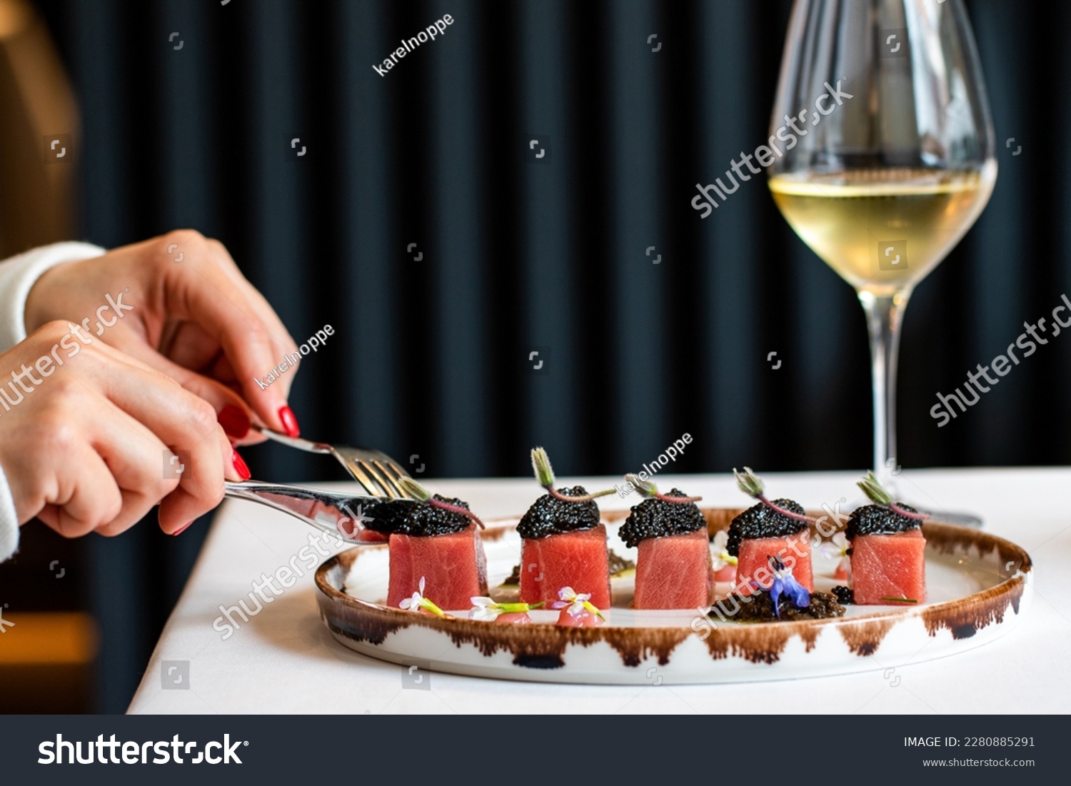 Close up detail of female having dinner in top notch restaurant. Side view of female hands next to blue fin tuna dish with beluga caviar and a glass of white wine. #2280885291
