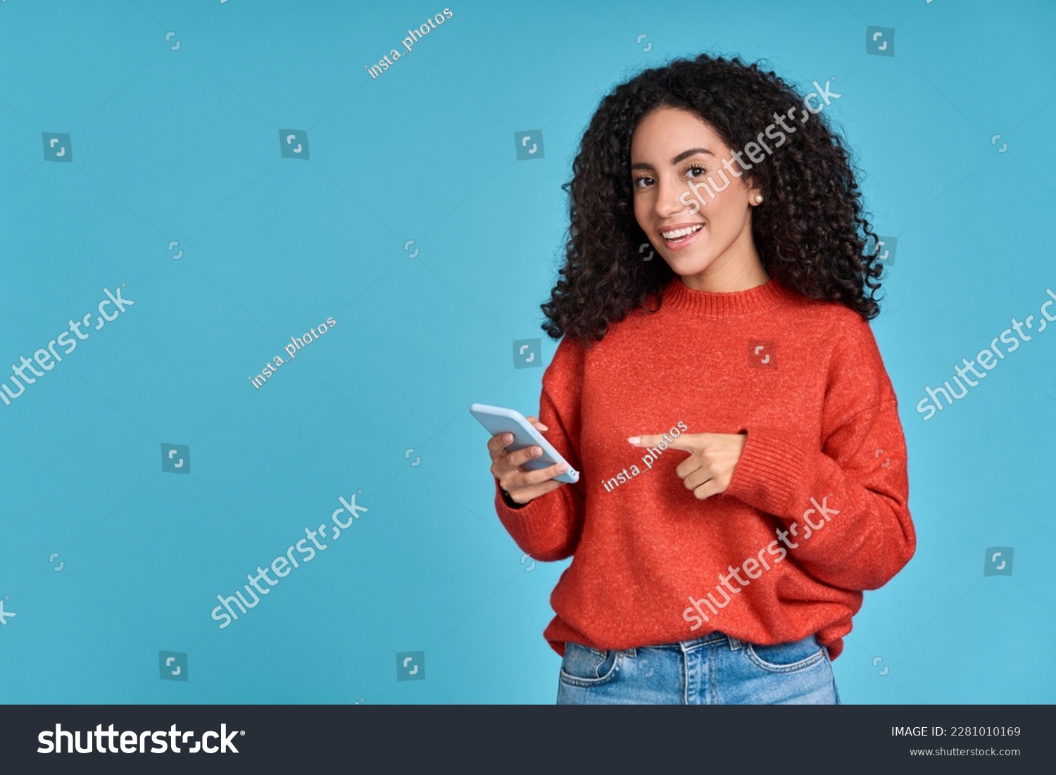 Young happy excited latin woman pointing at mobile phone isolated on blue background. Smiling female model holding cellphone using cell presenting advertising new trendy application concept. #2281010169
