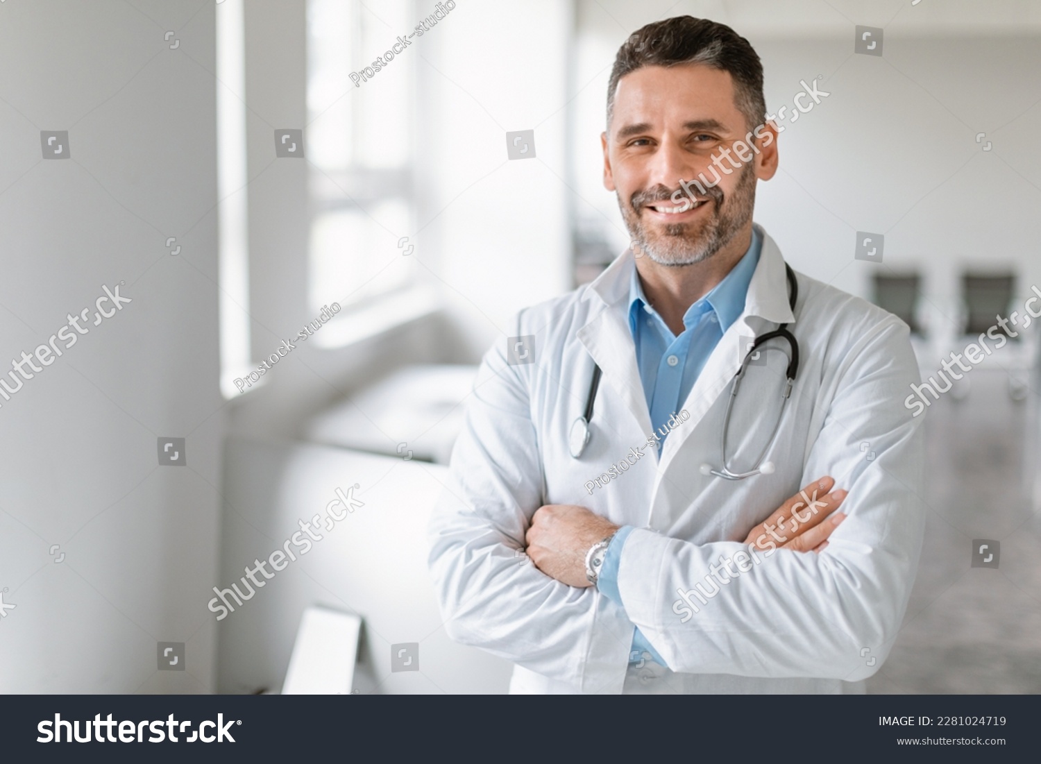 Portrait of friendly middle aged european male doctor in workwear with stethoscope on neck posing with folded arms in clinic interior, looking and smiling at camera, free space #2281024719
