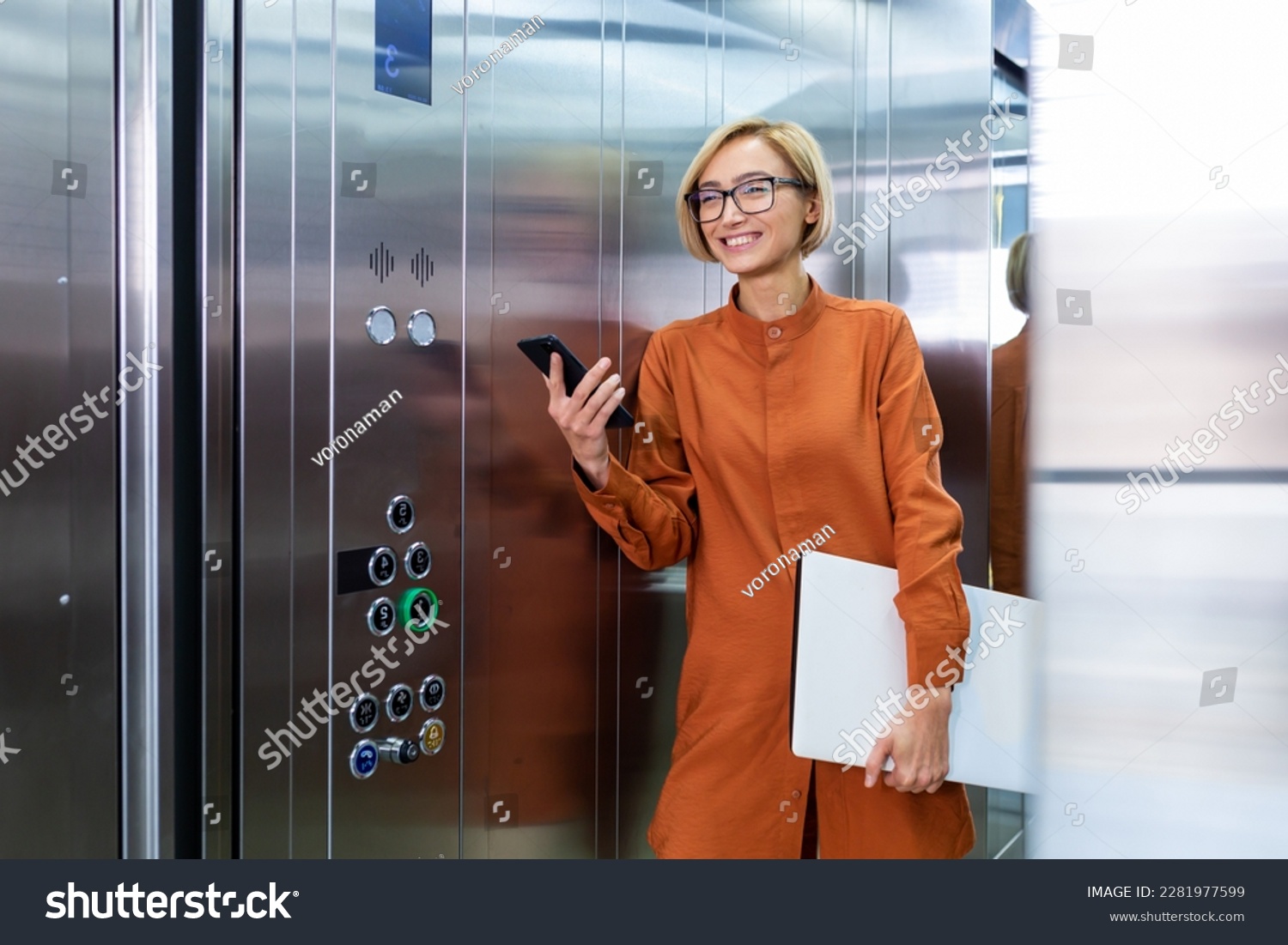 A smiling young business woman is standing in the office center in the elevator, holding a folder with documents and a phone in her hands, going to a conference. meeting. #2281977599