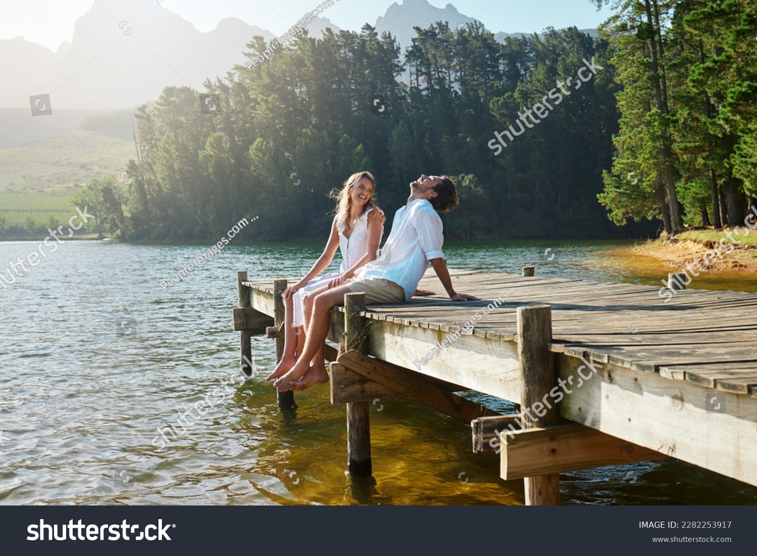 Young couple, lake and jetty with laughing, happiness and bonding with love in nature for holiday. Man, woman and comic joke on bridge by water to relax with conversation, care and vacation in summer #2282253917